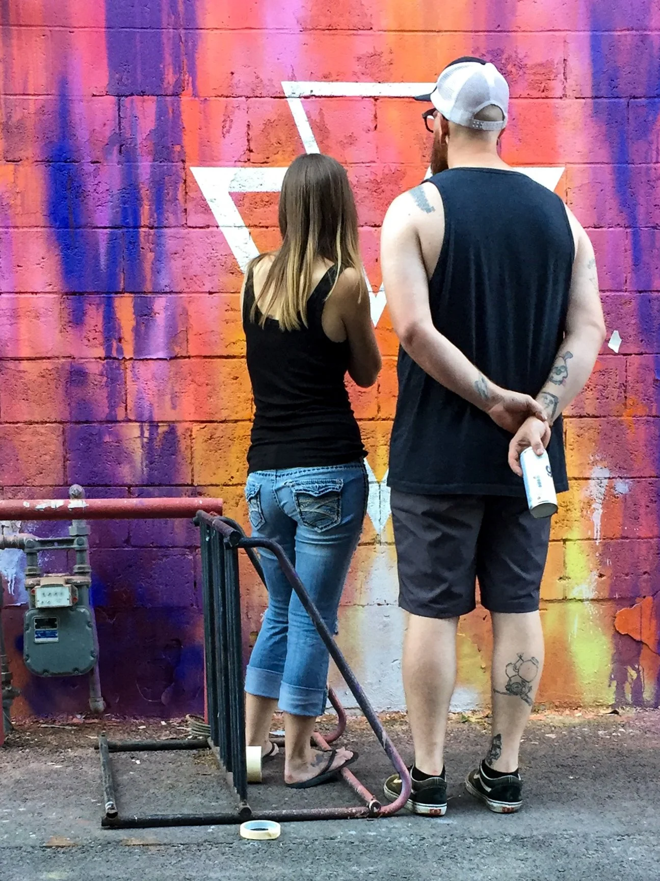 Two young people, a woman and a man, stand with their backs facing a colorful graffiti wall. The woman wears a black tank top and ripped jeans, and the man wears a sleeveless black shirt, shorts, a cap, and has tattoos on his arms and legs. They are 