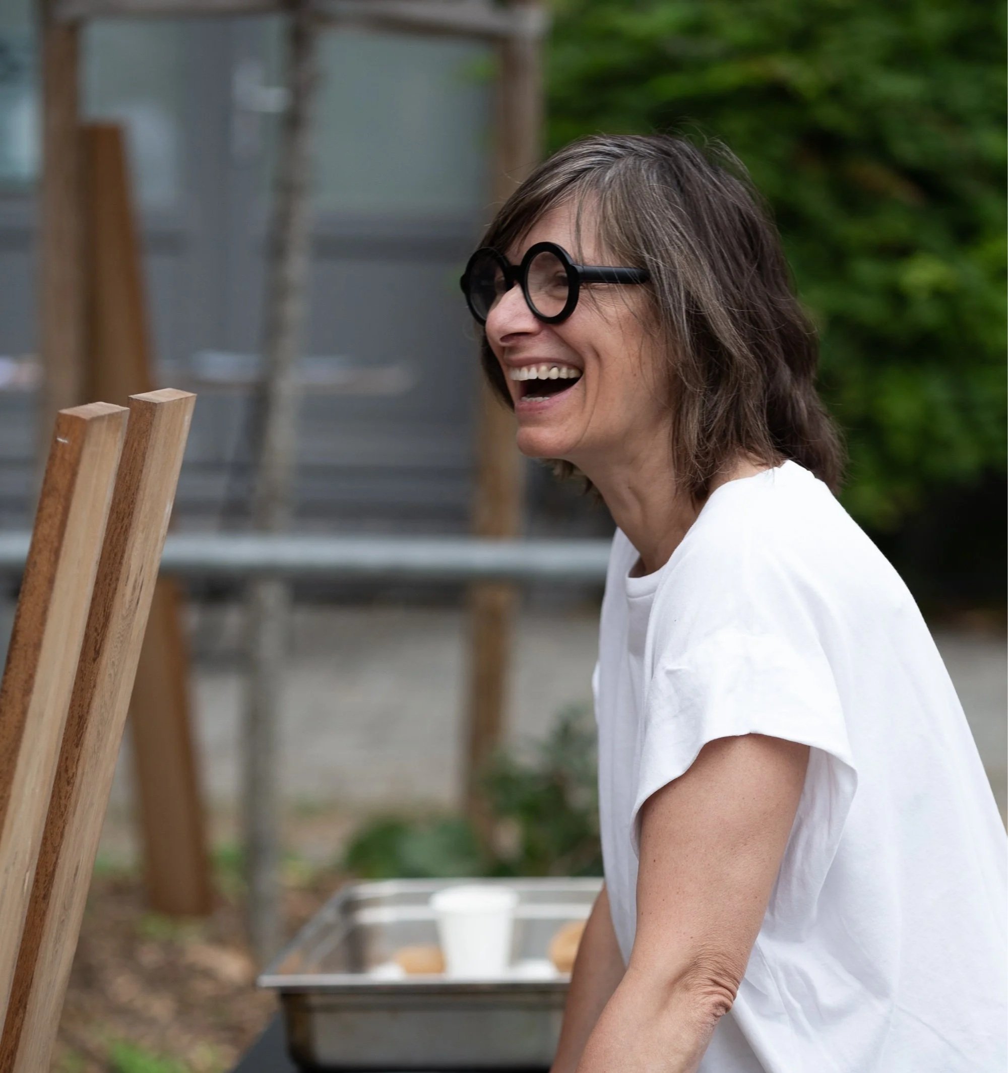 A woman with wavy brown hair and wearing black glasses, smiling, standing outdoors near a wooden structure with green foliage in the background.