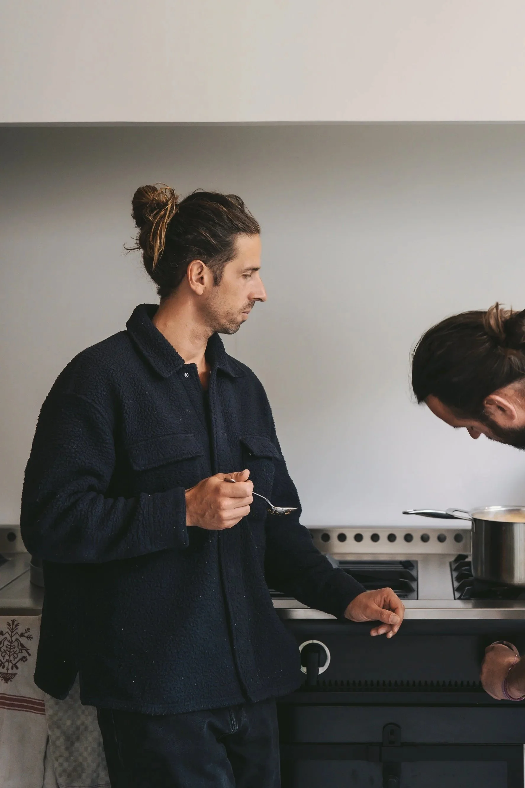 A man with long hair tied in a bun, wearing a dark fleece jacket, holding a spoon, standing in a kitchen near a stove, with a pot on the stove.