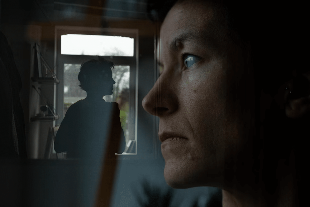 A close-up of a woman's face looking pensively to the right, with a silhouette of a person standing in front of a window in the background.