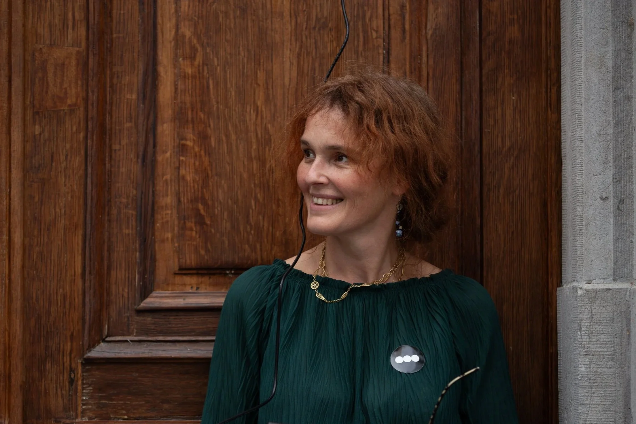 Woman with curly red hair smiling, wearing earrings, gold necklace, and green dress, standing in front of a wooden background.