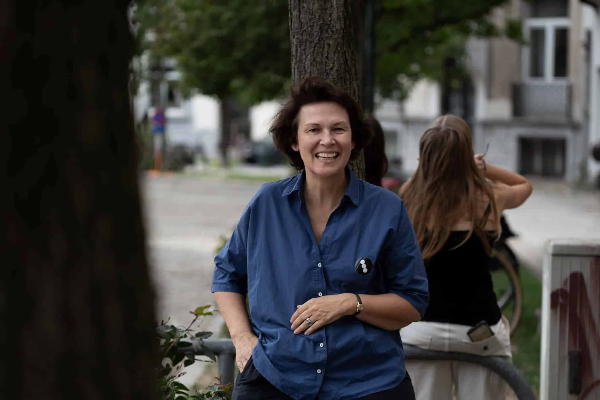 A woman with short dark hair smiling, wearing a blue button-up shirt, standing outdoors near a tree with buildings in the background.