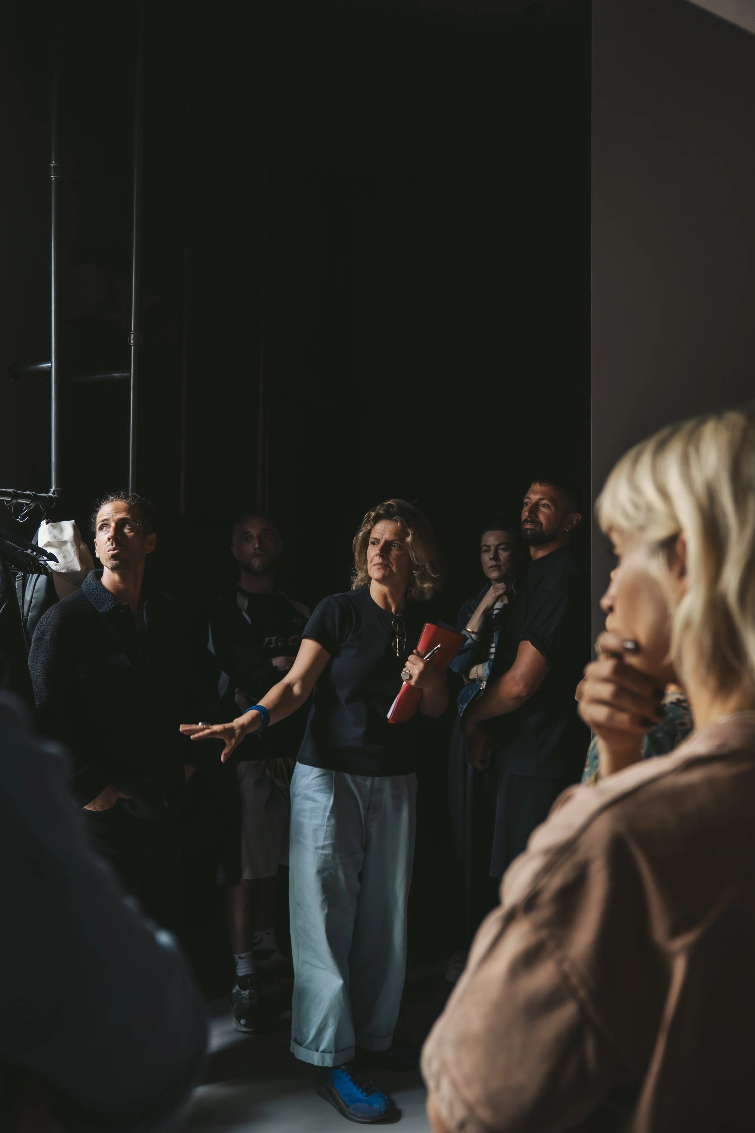 A group of people standing in a dark room, some appearing to be engaged in conversation or listening to someone outside of the frame. One woman in the center is holding a red notebook and gesturing with her hand.