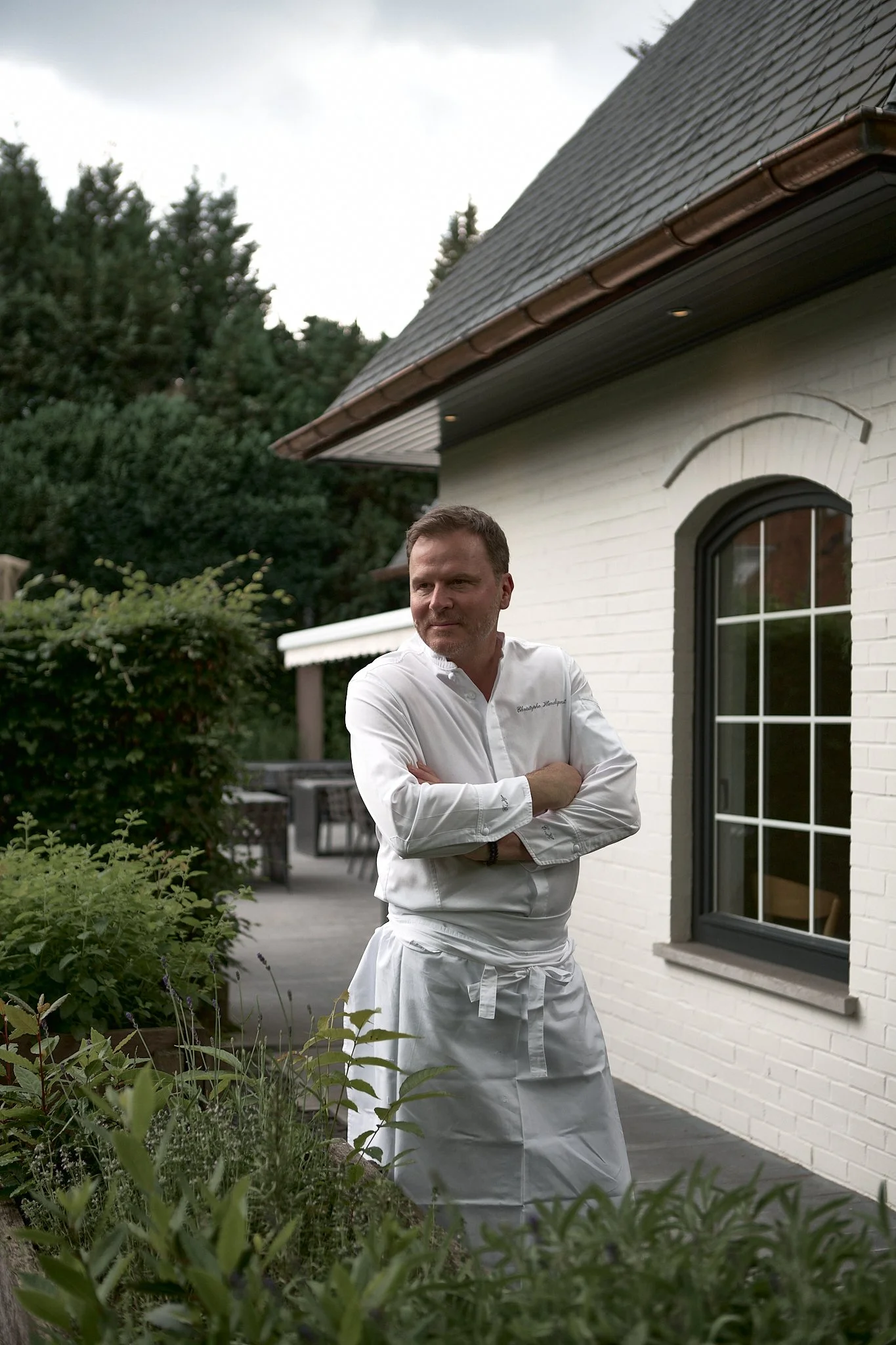 A man in a white chef's uniform standing with arms crossed outside a white brick house, surrounded by greenery and plants.