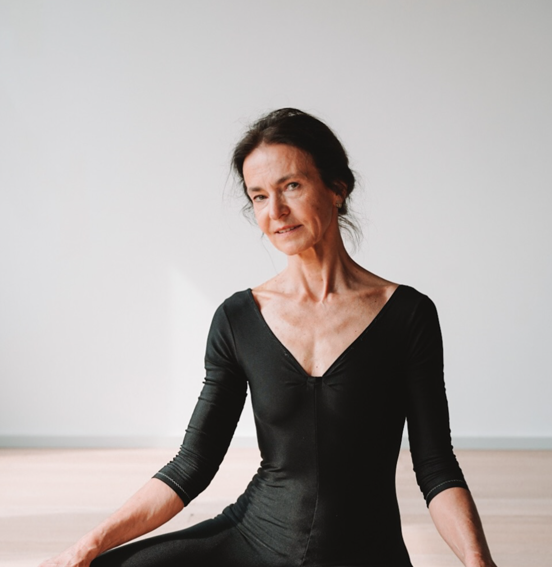 A woman practicing yoga or meditation indoors, sitting cross-legged on a light-colored wooden floor against a white wall, wearing a black long-sleeved top.