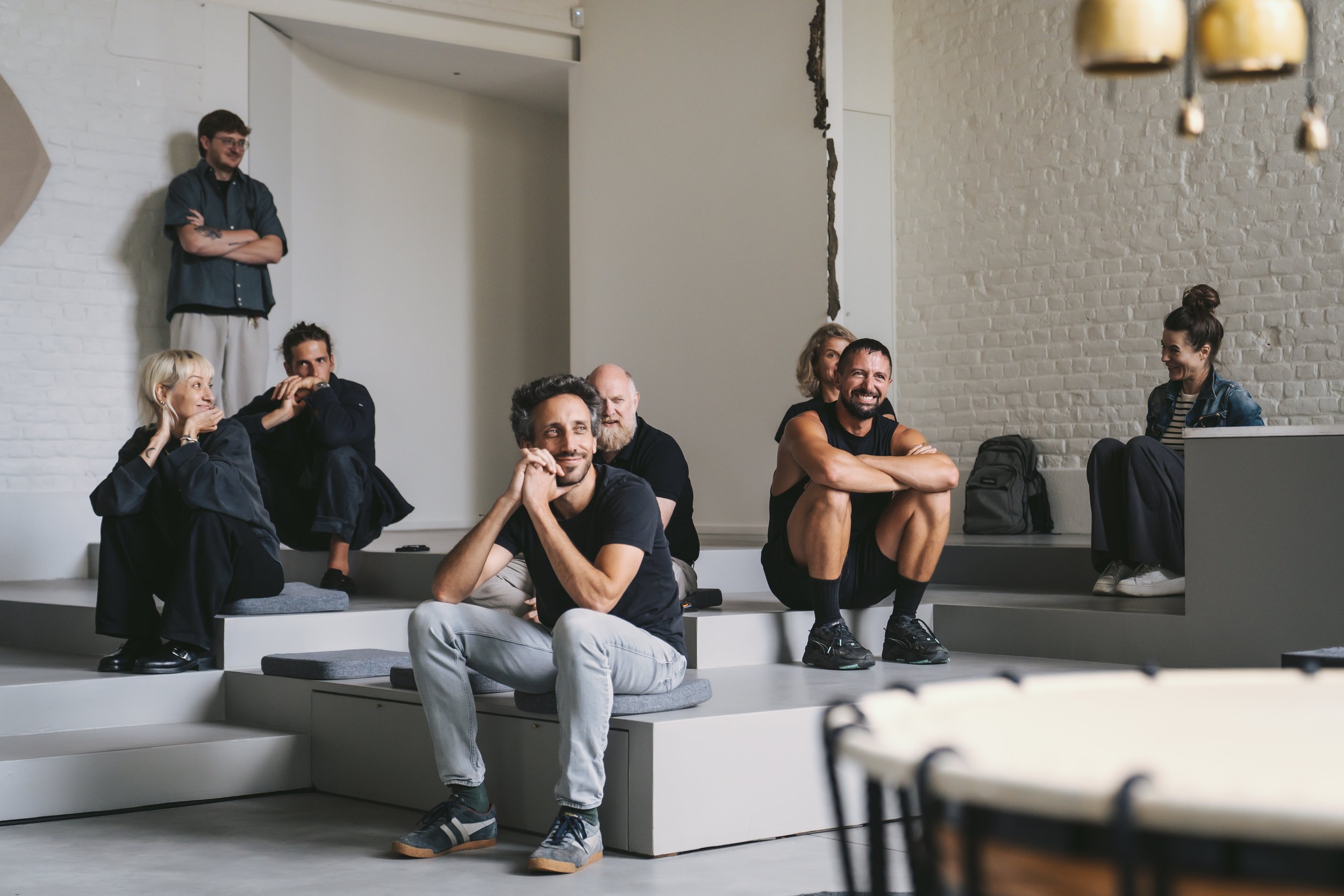 People sitting and standing on a tiered seating area in a modern, white-walled room, some smiling and engaging in conversation, with a focus on a smiling man in a black shirt sitting cross-legged in the front.