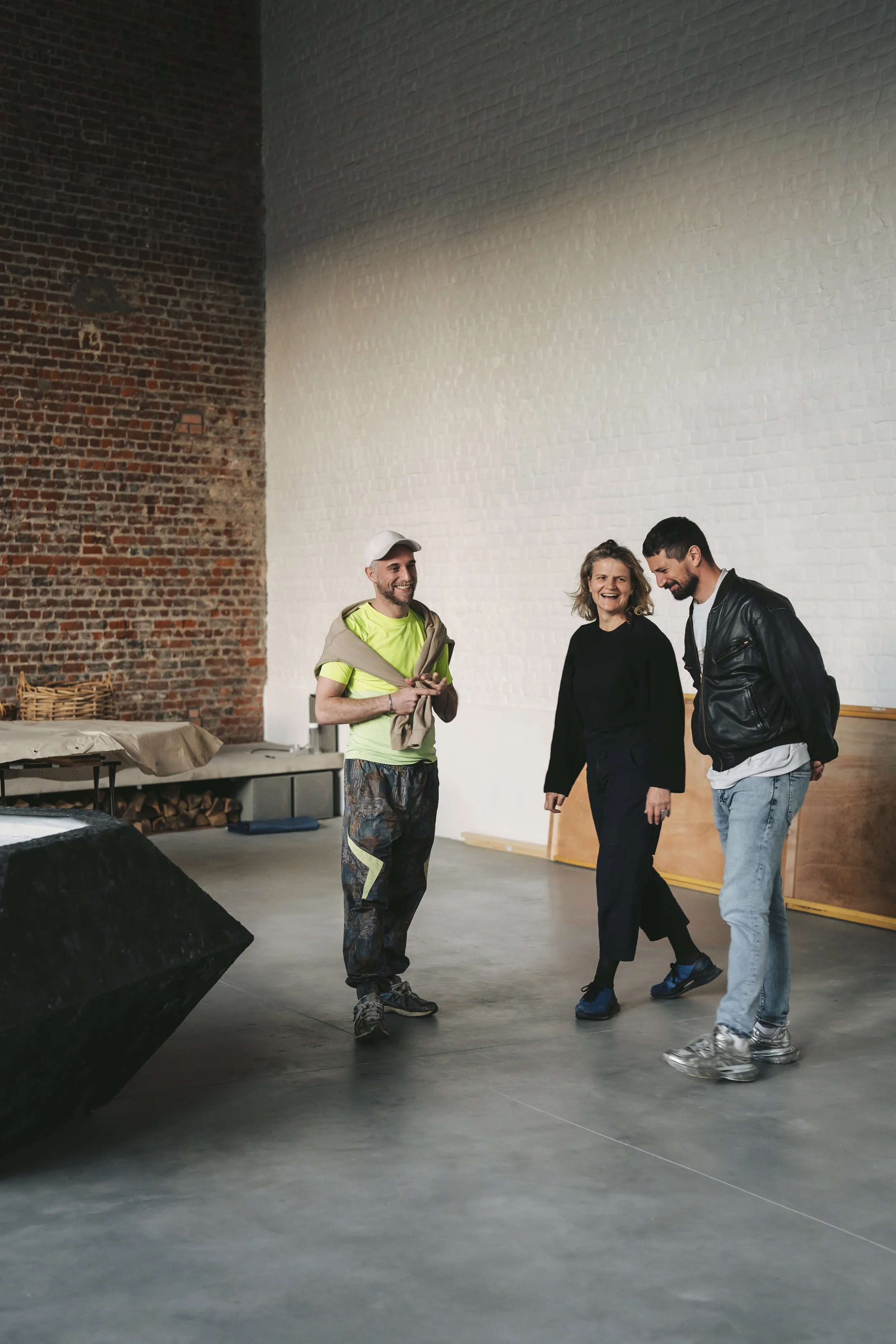 Three people standing and talking in an industrial-style room with brick and white walls, a table with some items, and a large black speaker.