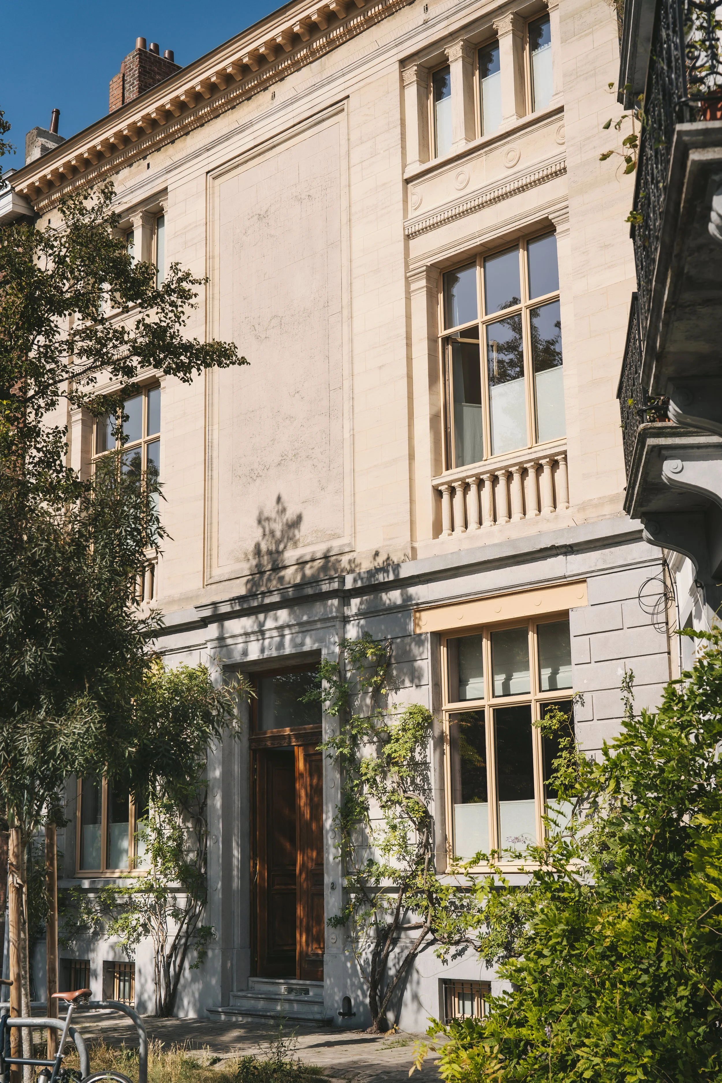 A multi-story beige stone building with large windows and decorative architectural elements, surrounded by trees and greenery, under a clear blue sky.