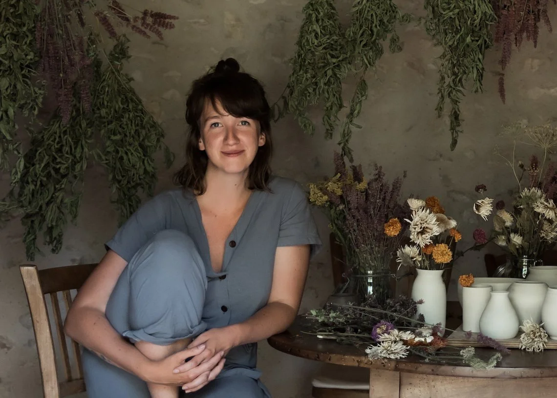 Young woman with dark brown hair, wearing a gray button-up shirt and light gray pants, sitting on a wooden chair in front of filled vases and dried flowers, with hanging greenery behind her.