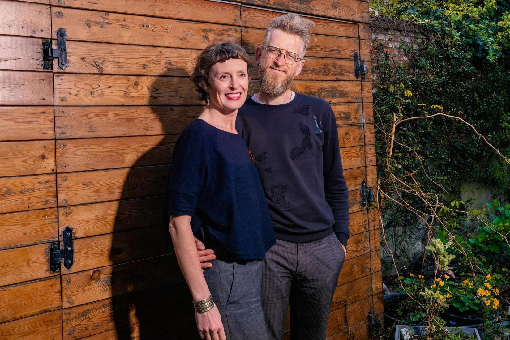 A woman and a man standing together outdoors in front of a wooden wall. The woman has short, curly hair, is wearing glasses, a dark blue top, and gray pants, and is smiling. The man has a beard, glasses, and styled hair, and is wearing a black sweater with patches and dark pants, with a slight smile. Bright sunlight casts shadows on the wall behind them, and there are some plants and greenery on the right side of the image.