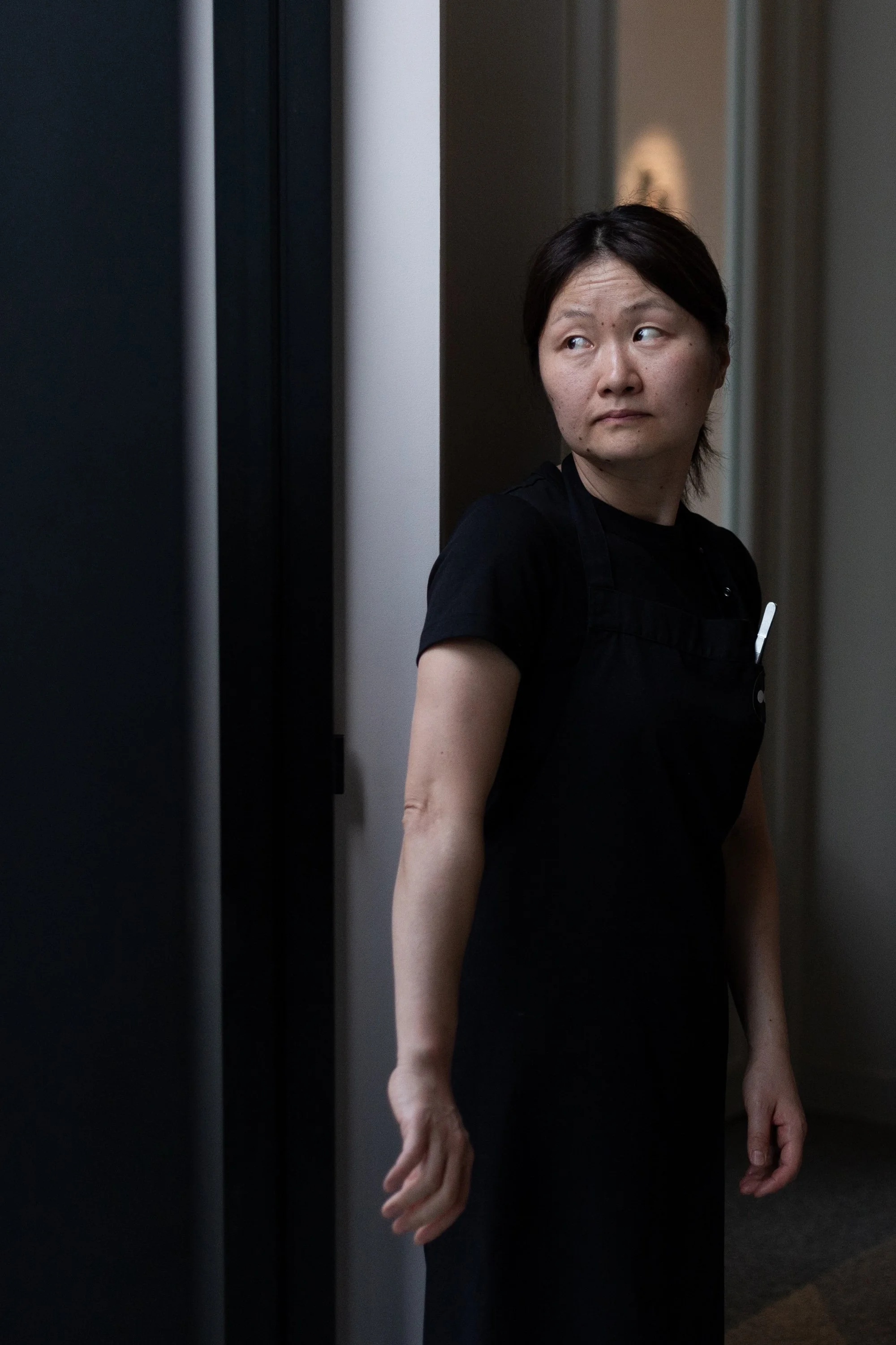 A woman with short dark hair and freckles is standing in an elevator, looking to her right. She is wearing a black shirt and has a pen tucked into her shirt pocket. The elevator wall is dark with light from behind, and her arm is down by her side.