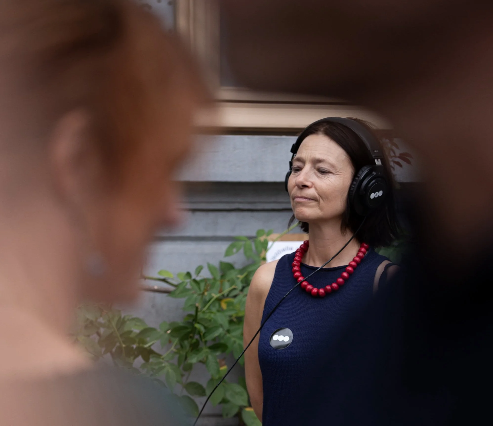 A woman with dark hair, wearing a navy sleeveless top, red beaded necklace, and large black headphones, is smiling with her eyes closed in front of a gray wall with plants.