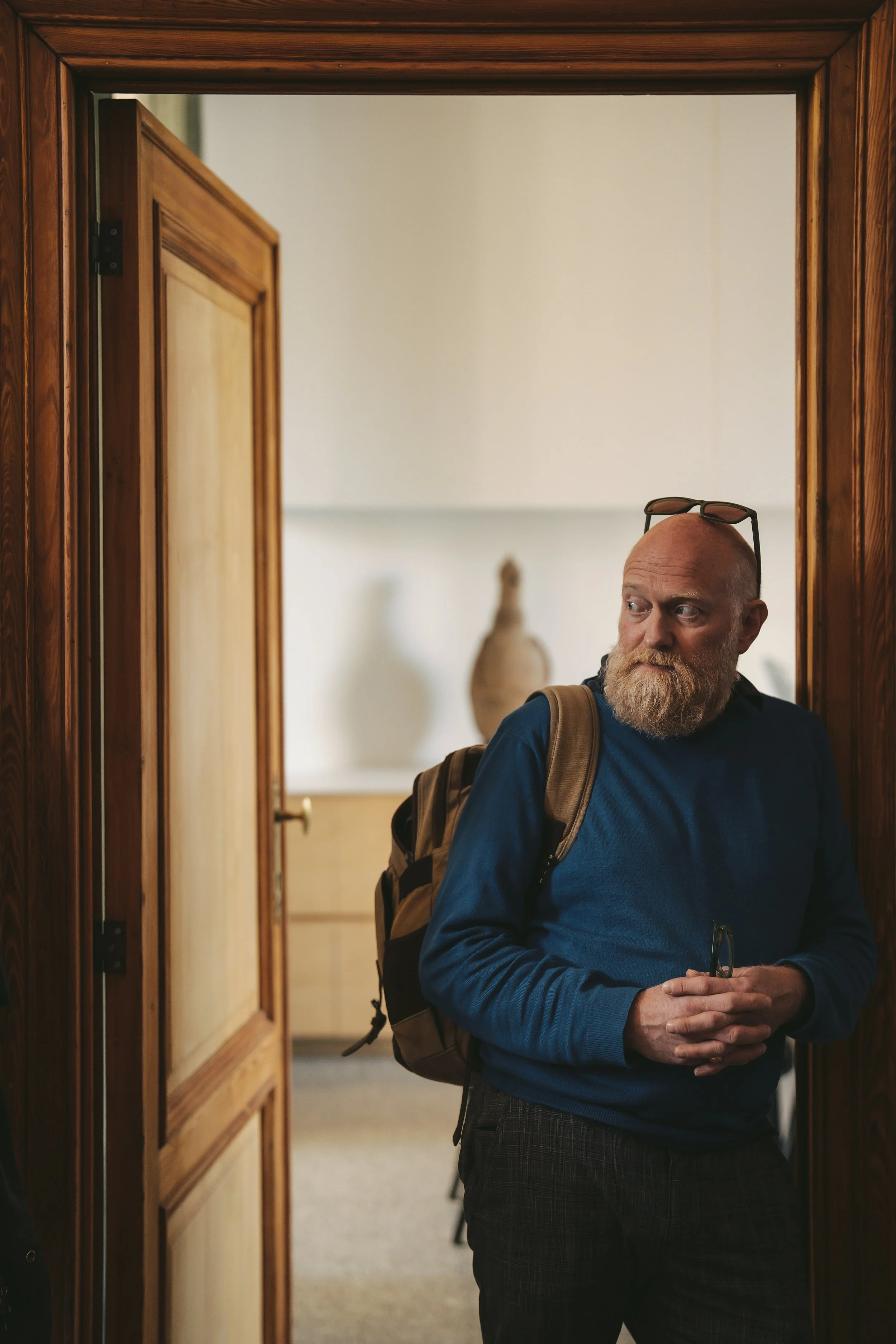 A man with a beard and glasses resting on his head, wearing a dark blue sweater and carrying a backpack, stands by a wooden doorframe, looking to his right, while holding sunglasses in his hands.