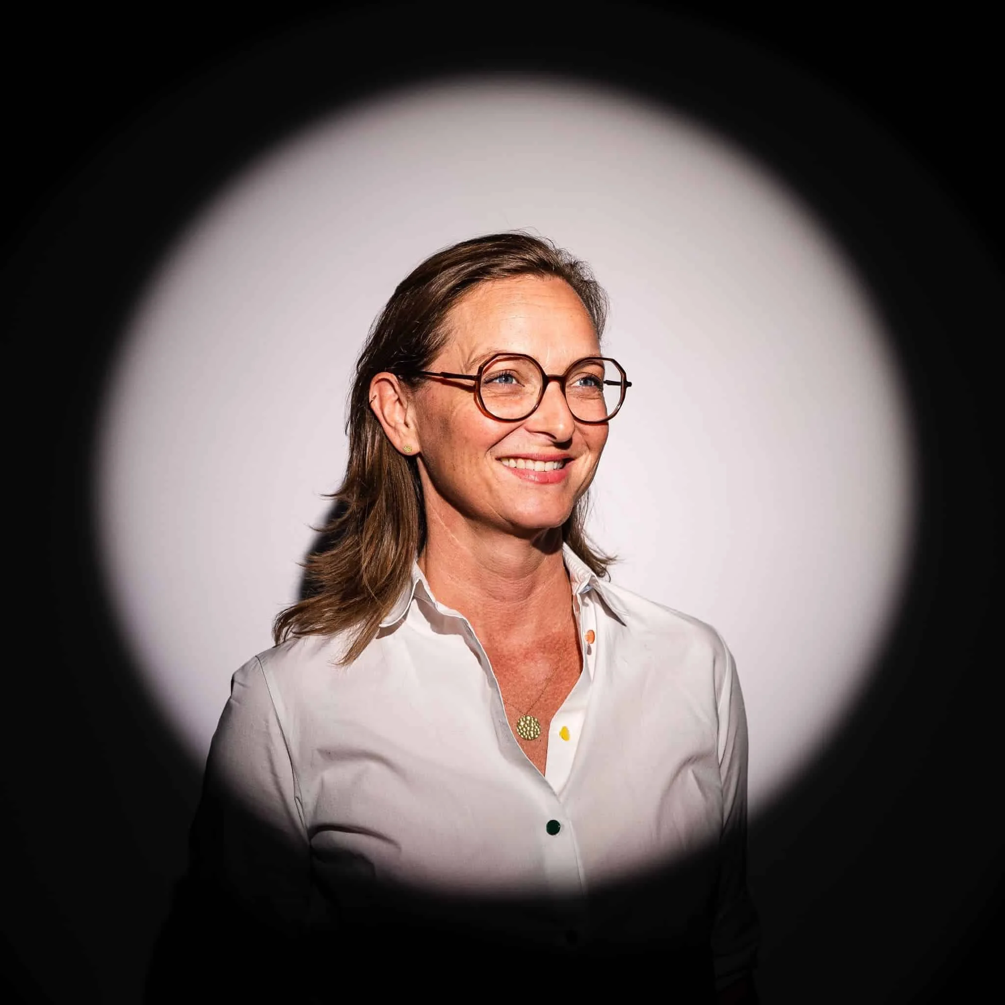 Smiling woman with glasses and earrings, wearing a white shirt with colorful buttons, against a plain background.
