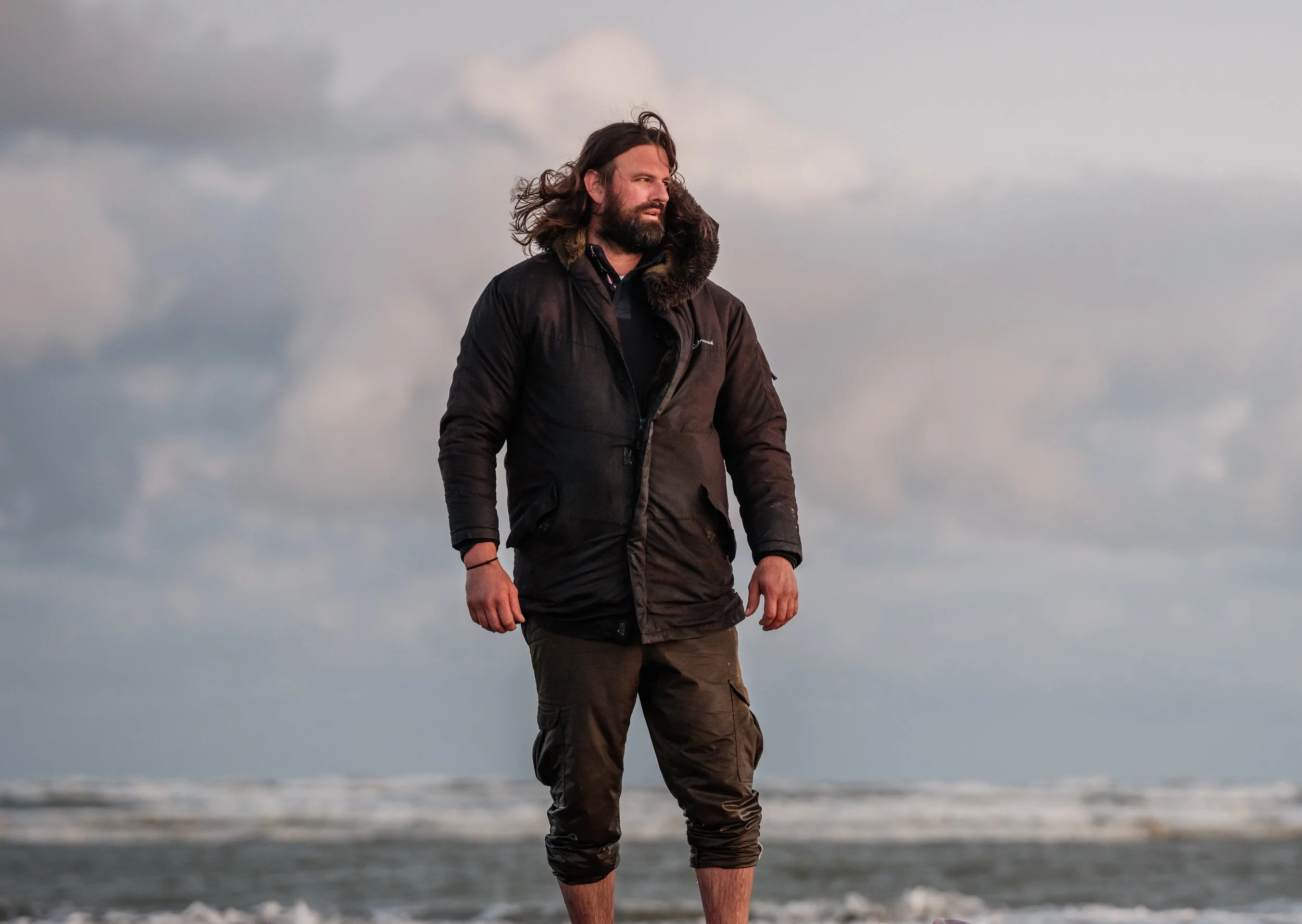 A man with long hair and a beard walking on the beach, wearing a black jacket and brown pants, with cloudy sky in the background.