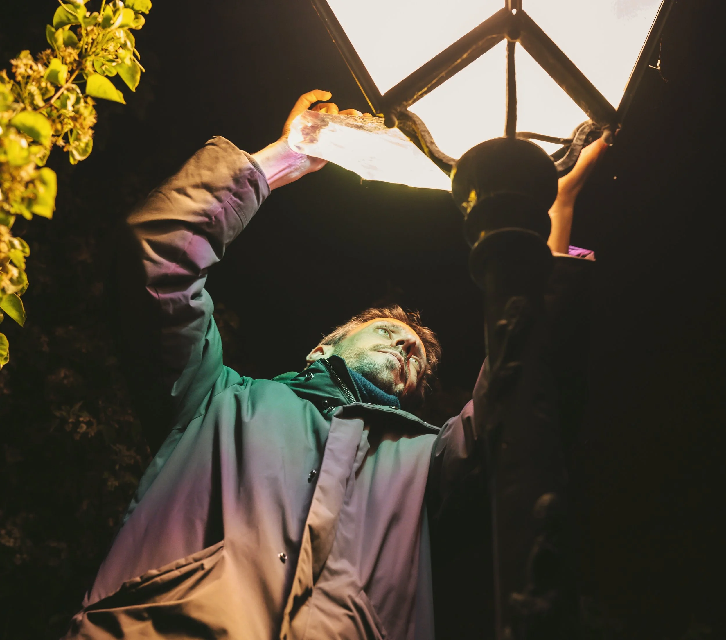 A person lying on the ground at night, gazing up at a bright outdoor light fixture with two lightbulbs and holding onto it.