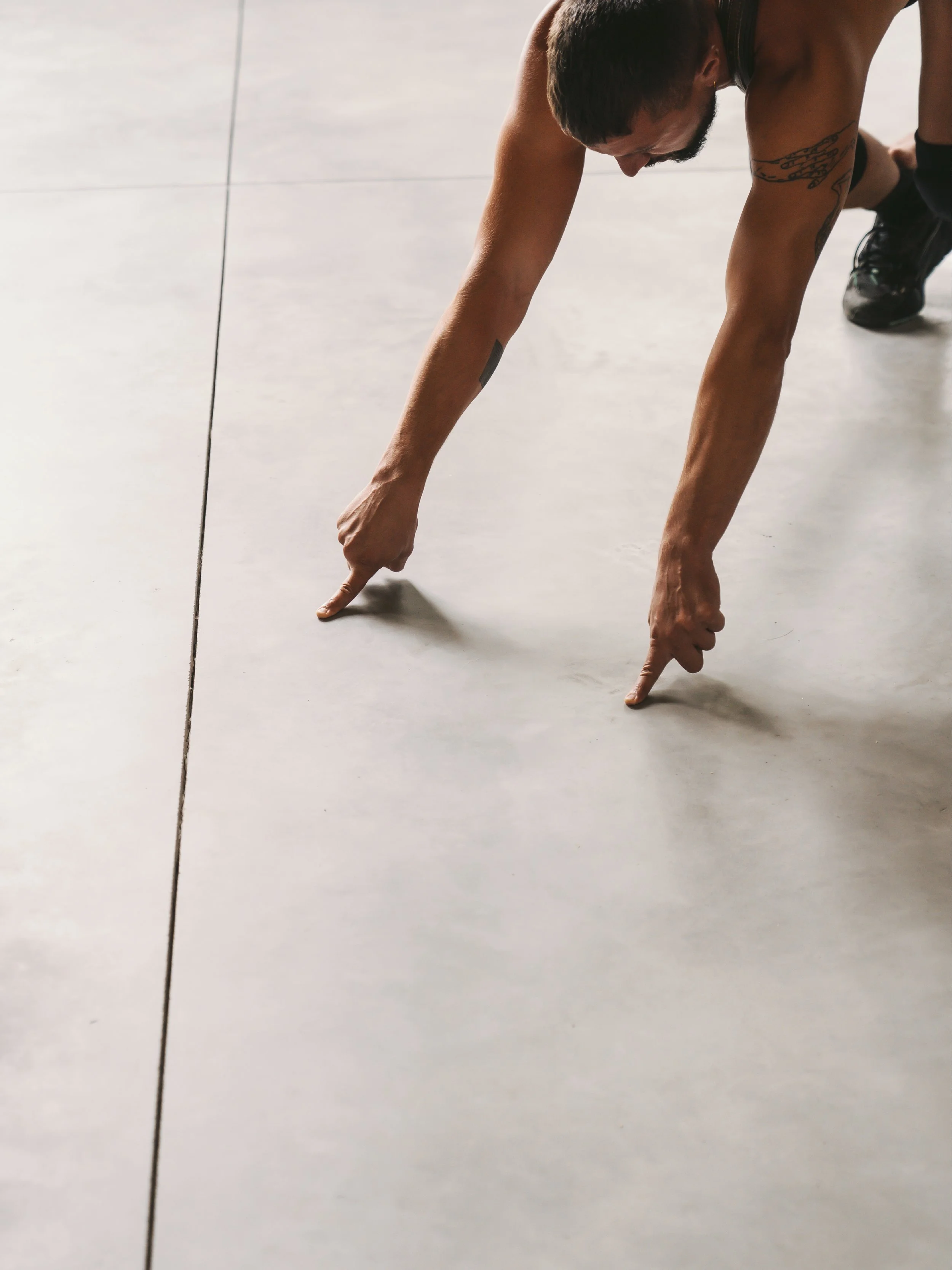 A man in athletic gear, with tattoos on his arms, is on all fours on a smooth floor, pointing with both index fingers toward the ground.