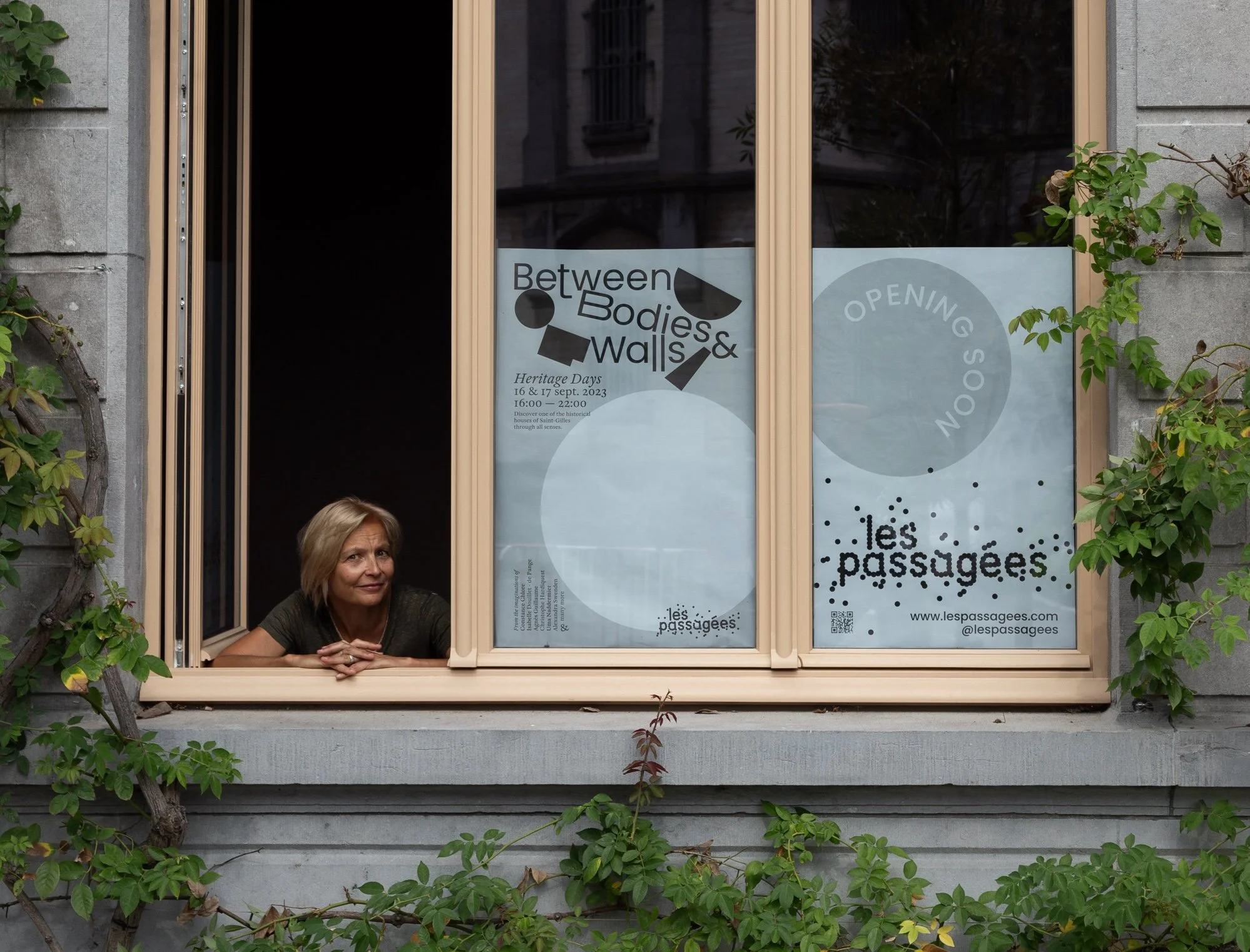 A woman with short blonde hair sitting at a window sill with a smile. The window has signs for an event called "Between Bodies & Walls" on September 16 and 17, 2023, with an open session indicated. Surrounding the window are green leafy plants climbing the wall.