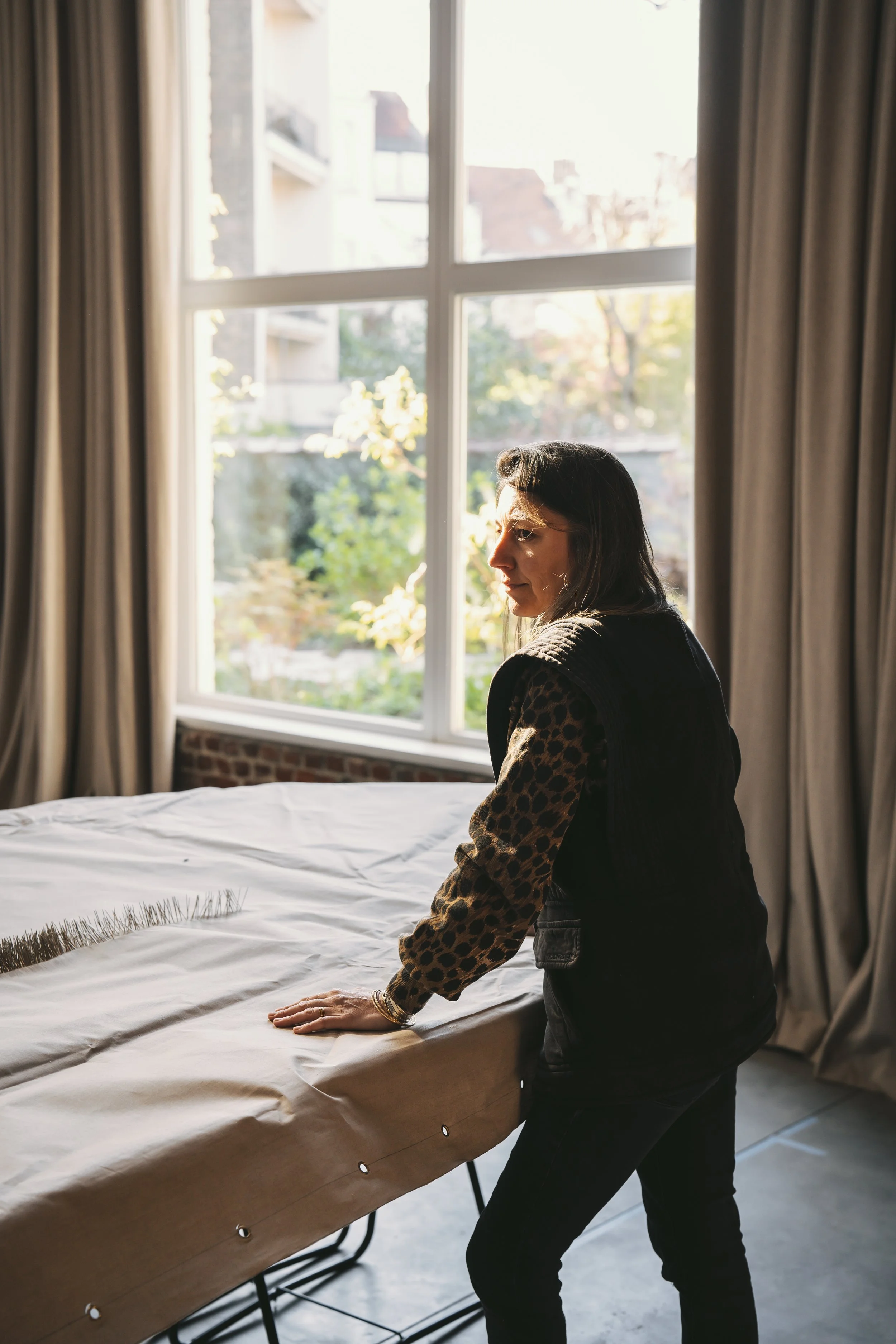 A woman in black jeans and leopard print long-sleeve shirt makes a bed in a bedroom with large window with beige curtains, sunlight streaming in, outside trees and buildings visible.