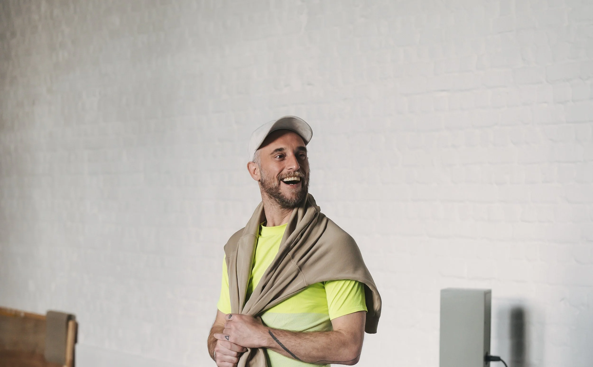 A man with a beard, wearing a beige cap, a bright yellow shirt, and a light jacket draped over his shoulders, smiling and looking to his left against a white brick wall.