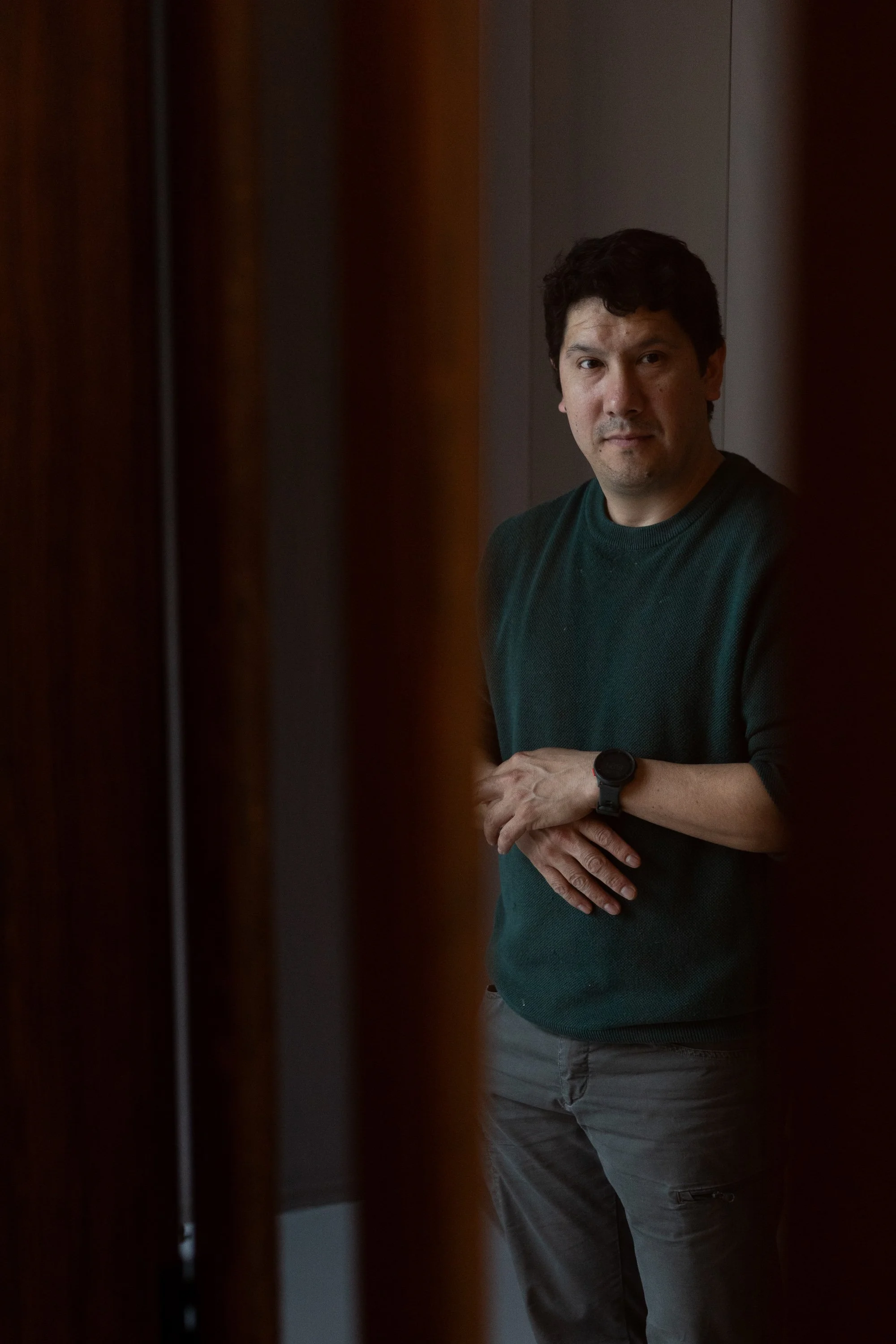 A man standing indoors, wearing a dark green sweater and gray pants, with a watch on his left wrist, partially framed by wooden slats.