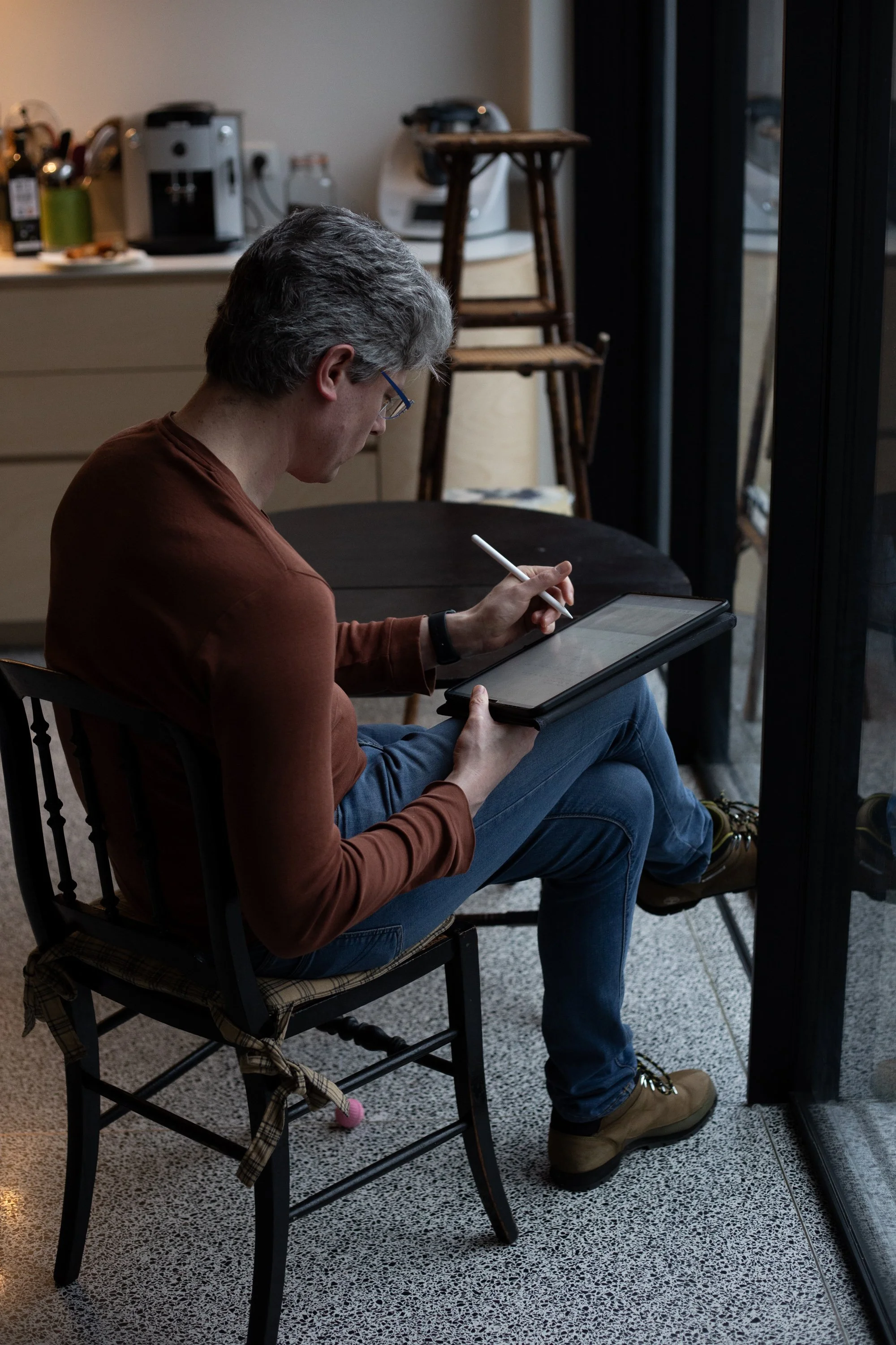 A person sitting on a wooden chair, wearing glasses and a brown long-sleeve shirt, using a stylus and tablet while sitting near a large glass window in a room with a kitchen in the background.