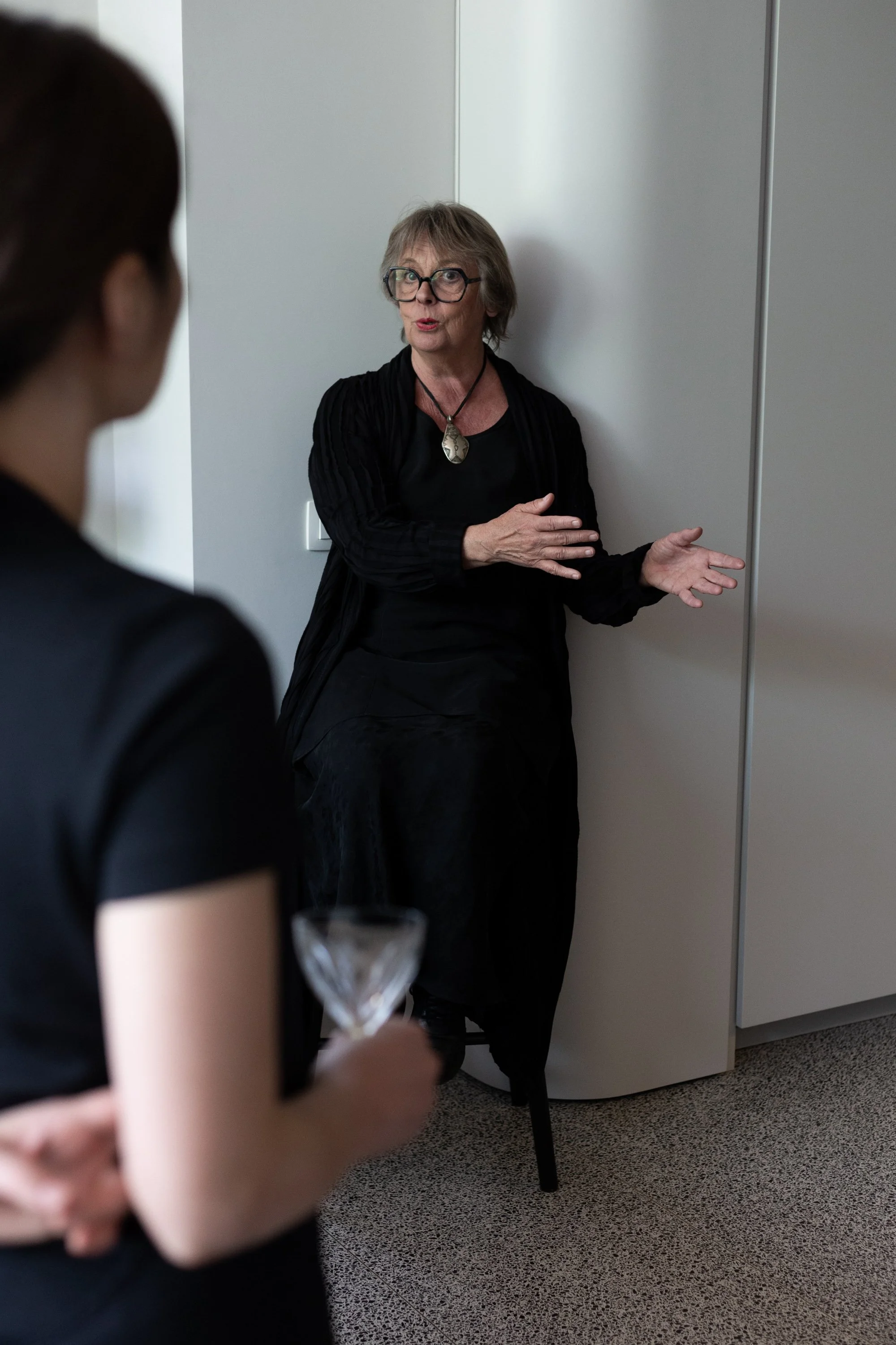 A woman with short gray hair, glasses, and a black dress stands near a wall, speaking or explaining to someone. In the foreground, a person is holding a glass and a drink. The setting appears to be an indoor event or gathering.