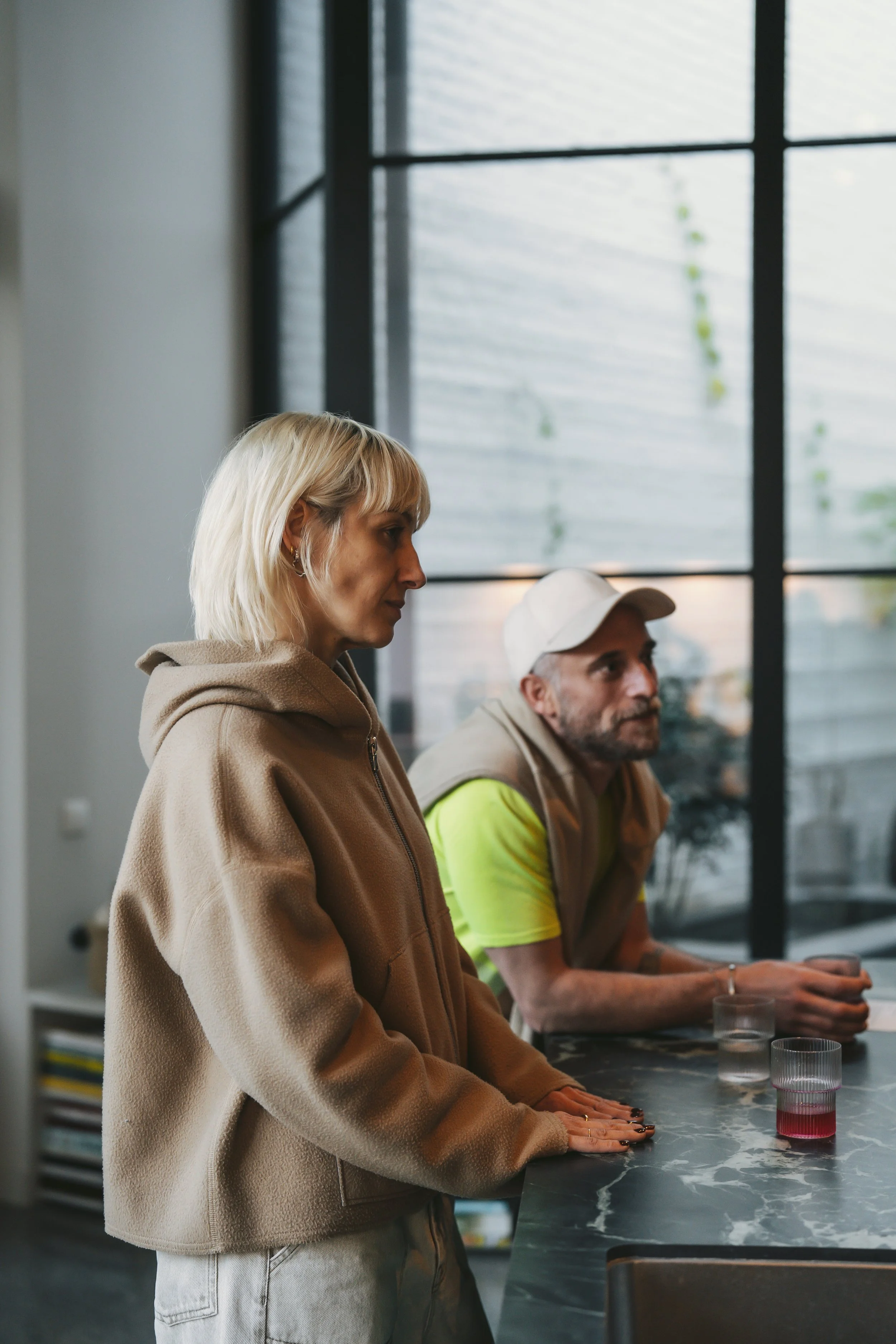 Two people, a woman and a man, are sitting at a table with their hands resting on the surface. The woman has shoulder-length blonde hair and is wearing a beige hoodie, while the man has a beard, is wearing a white cap, and a neon yellow shirt with a beige vest. They are indoors, with a large window behind them, and there are glasses with pink liquid on the table.