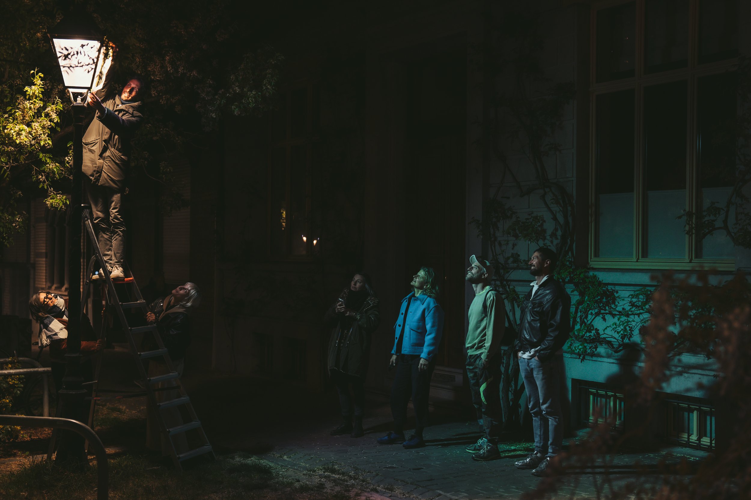A man on a ladder installing or adjusting a street lamp at night while a group of people stands nearby, looking at him. The background shows a dark, wooded area and a building with windows.