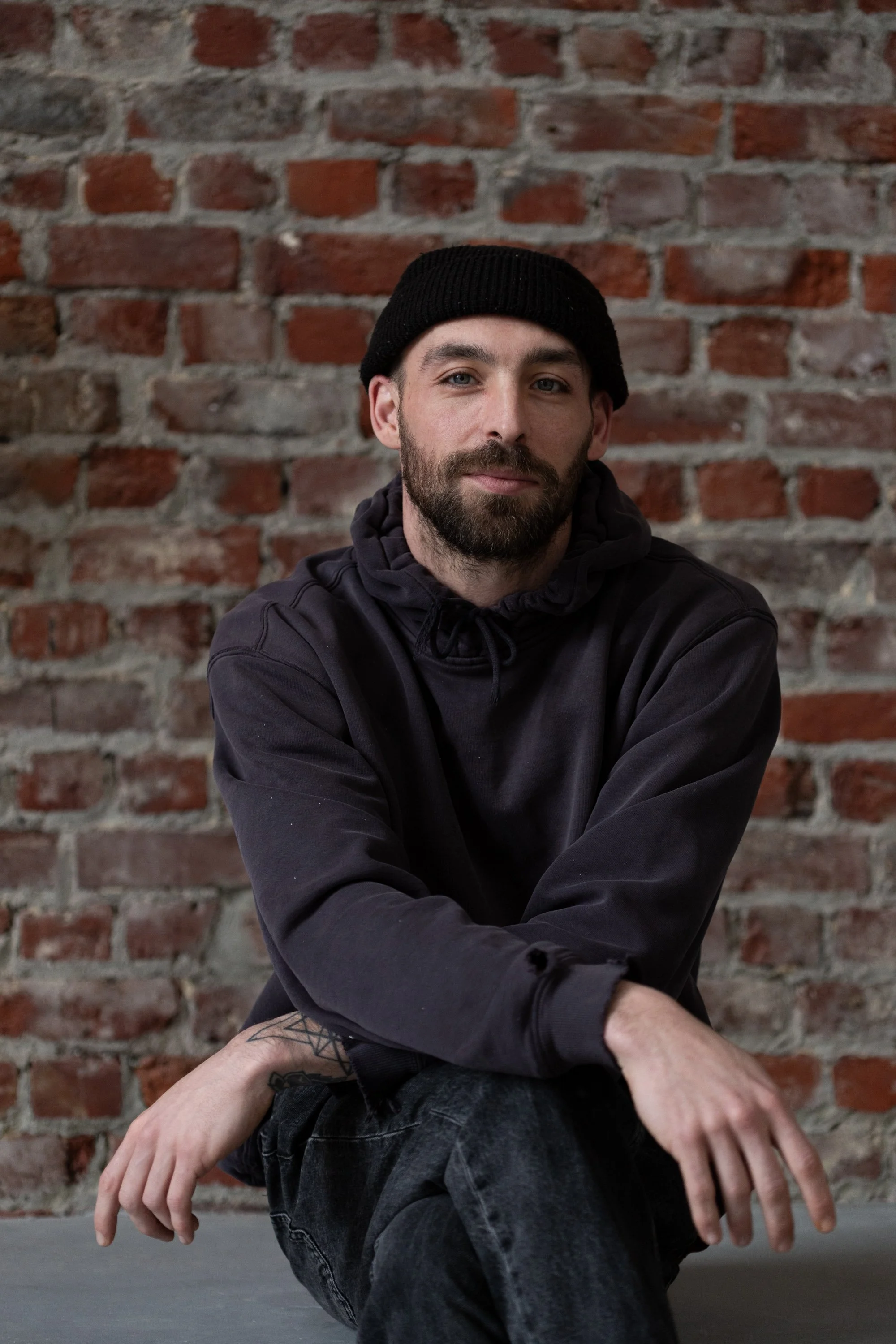 A man with a beard wearing a black beanie and black hoodie, sitting against a brick wall.