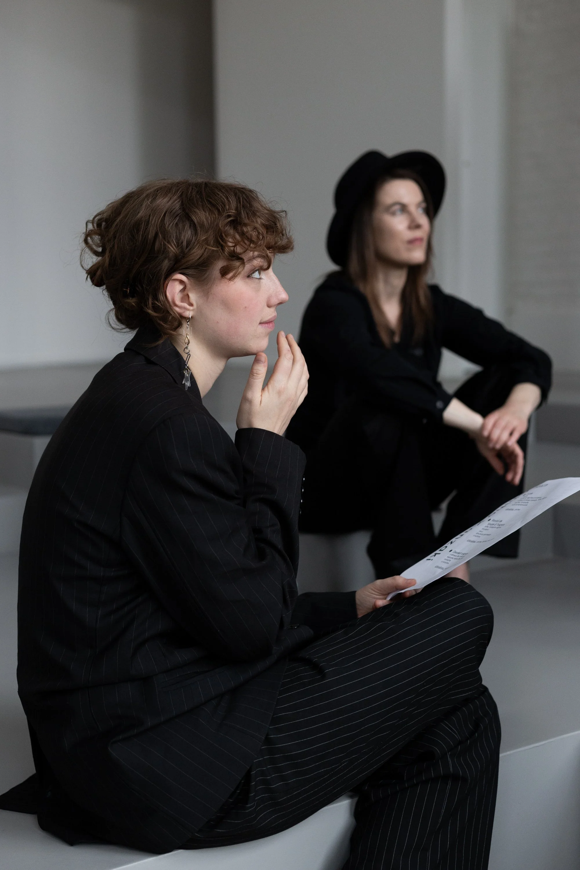 Two women sitting on a bench indoors, one with short curly hair looking to the side and holding a sheet of paper, the other with long straight hair and wearing a black hat, also looking to the side.