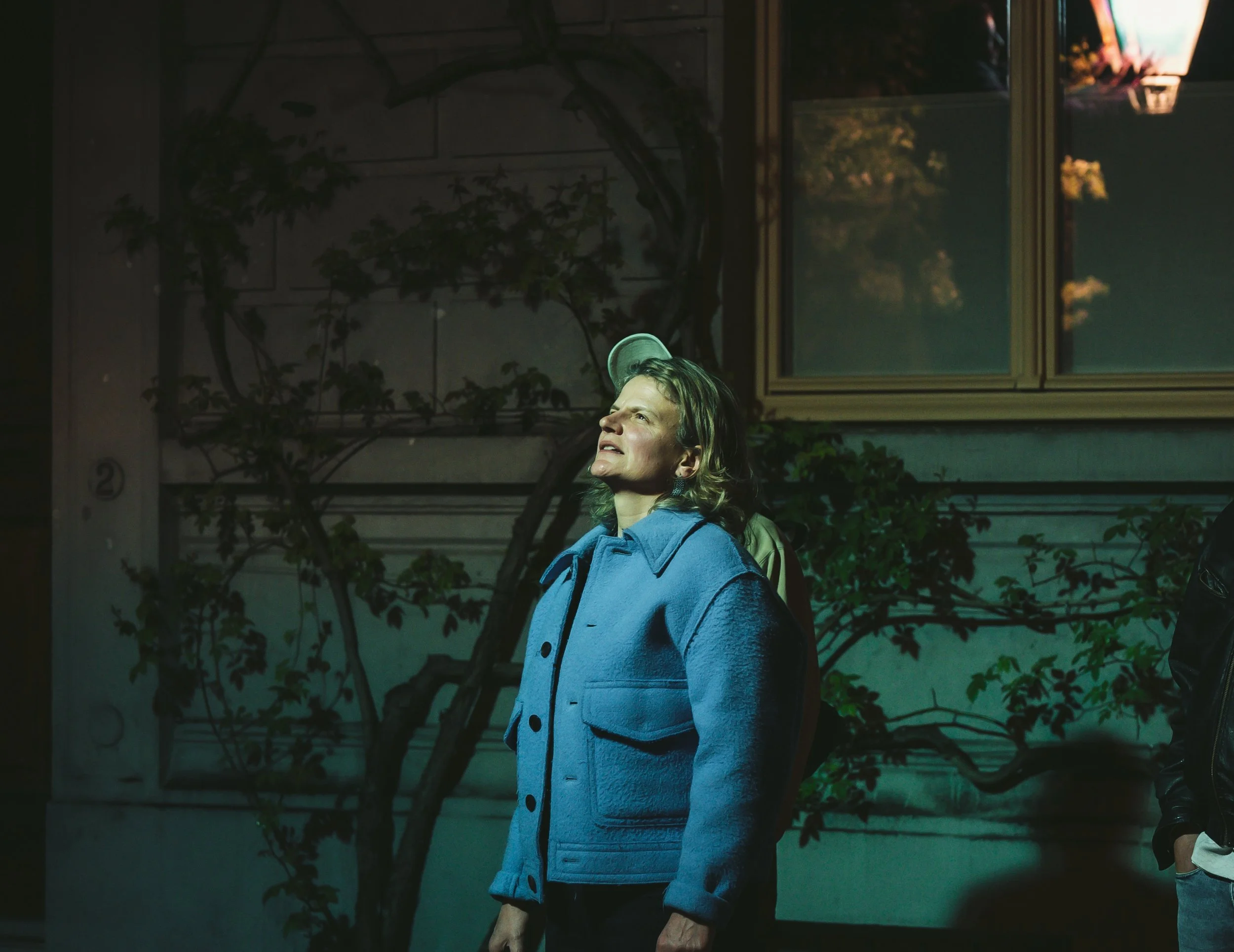 A woman with short hair wearing a blue jacket and a green cap, standing indoors near a large window and some plants.