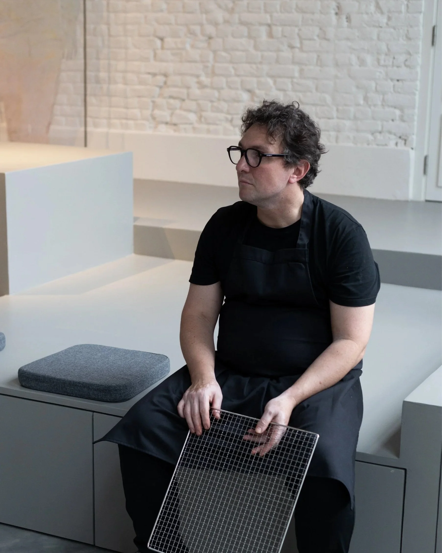 A man with curly hair, glasses, wearing a black shirt and apron, is sitting in a room with a white brick wall. He is holding a black and white grid-patterned object, possibly a cabinet or a screen. There is a gray counter or table with a gray cushion on top in front of him.