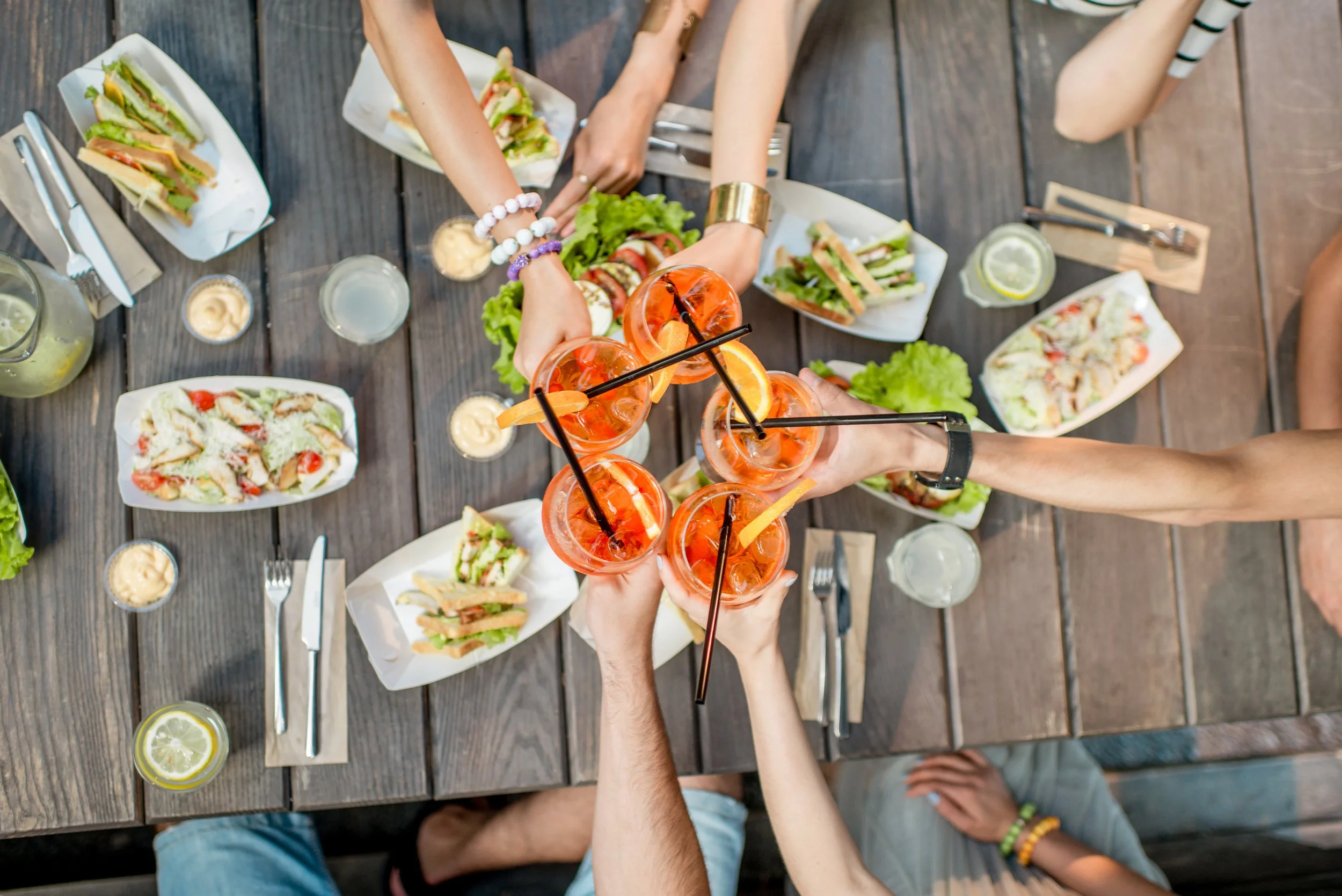 People clinking glasses with orange drinks garnished with orange slices at a table set with sandwiches, salads, and lemon water glasses.