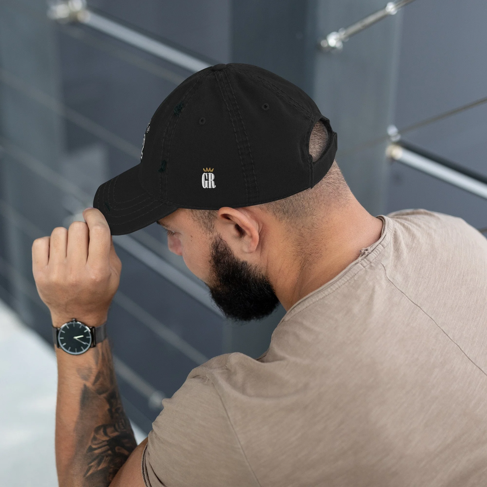 A man with a beard wearing a beige t-shirt and black baseball cap with the initials 'GR' in white and a small gold crown logo, taking down the cap from his head to reveal his face, with a metallic wall and ceiling pipes in the background.