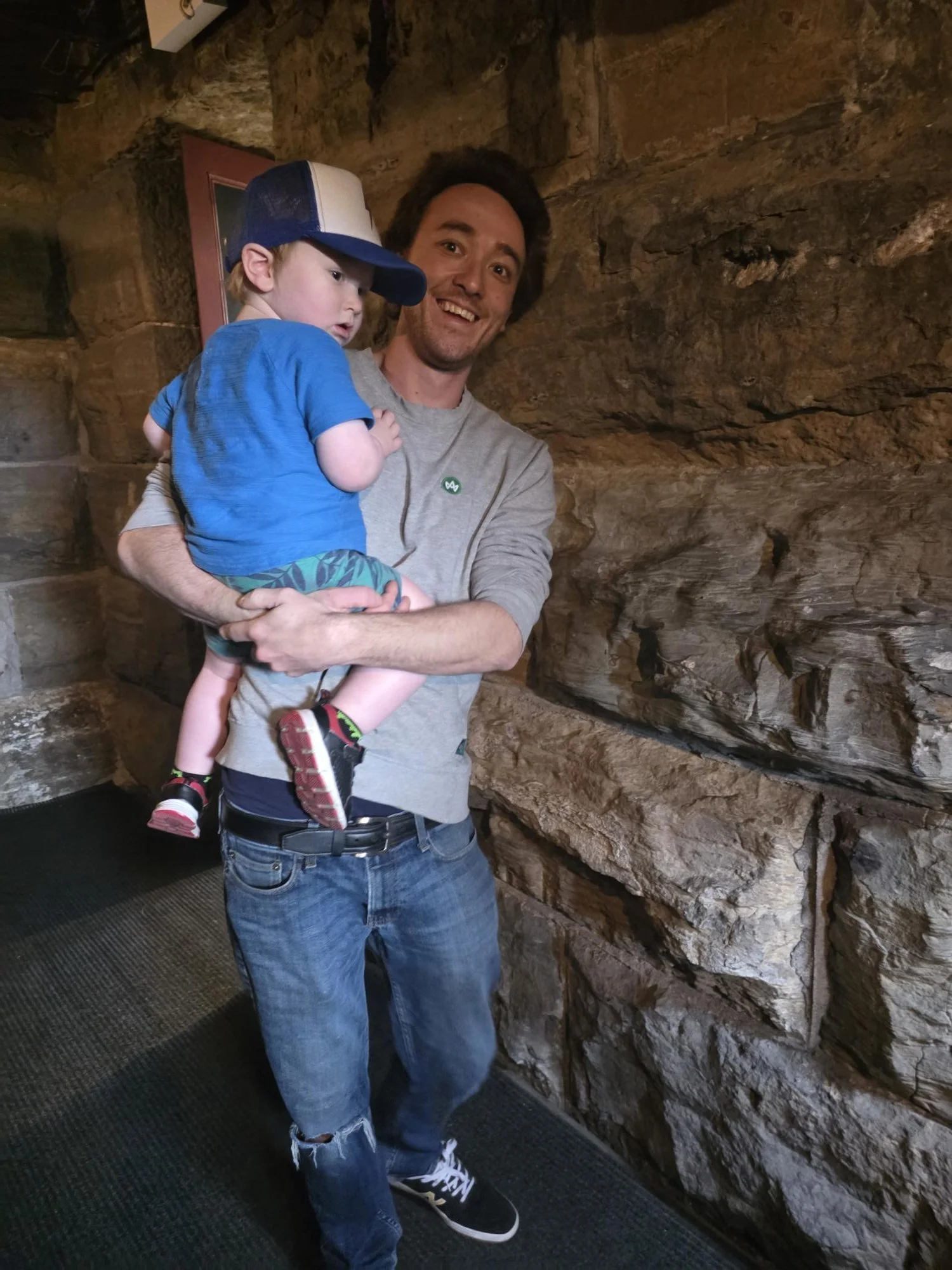 A man holding a young boy in front of a rocky wall inside a building.