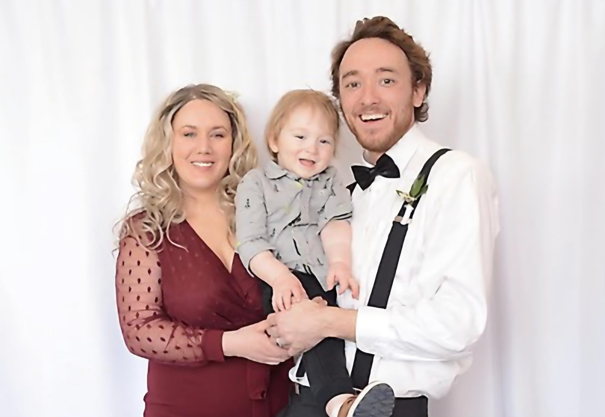 A family of three posing together against a white background. The woman has long blonde curly hair and is wearing a maroon dress with sheer polka-dot sleeves. The man has brown hair, a beard, and is dressed in a white tuxedo shirt with a black bow tie and suspenders. They are holding a young boy with red hair, who is wearing a gray shirt with black stripes and has a happy expression.