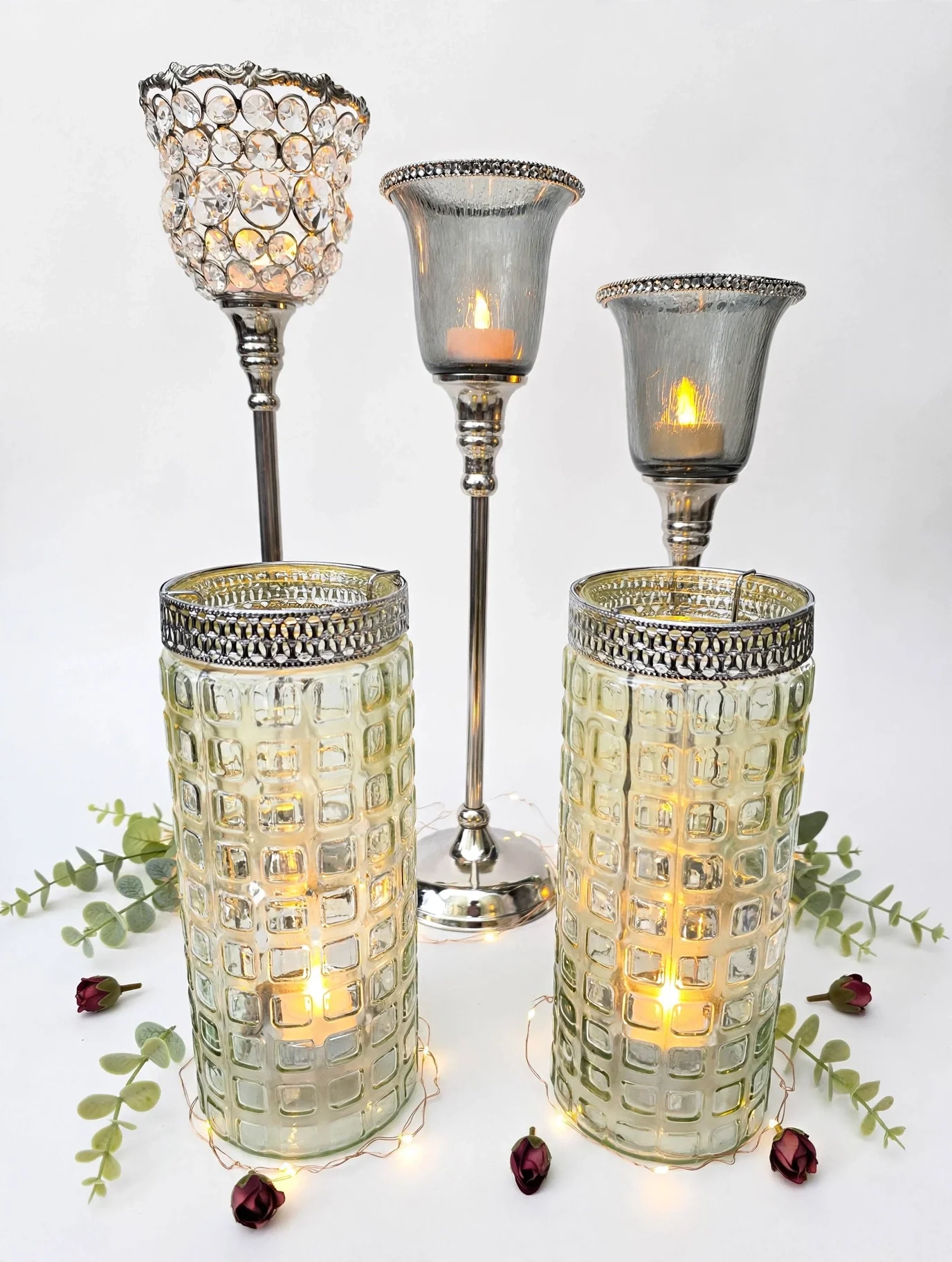 Silver and glass candle holders with lit candles, greenery, and small red rose accents