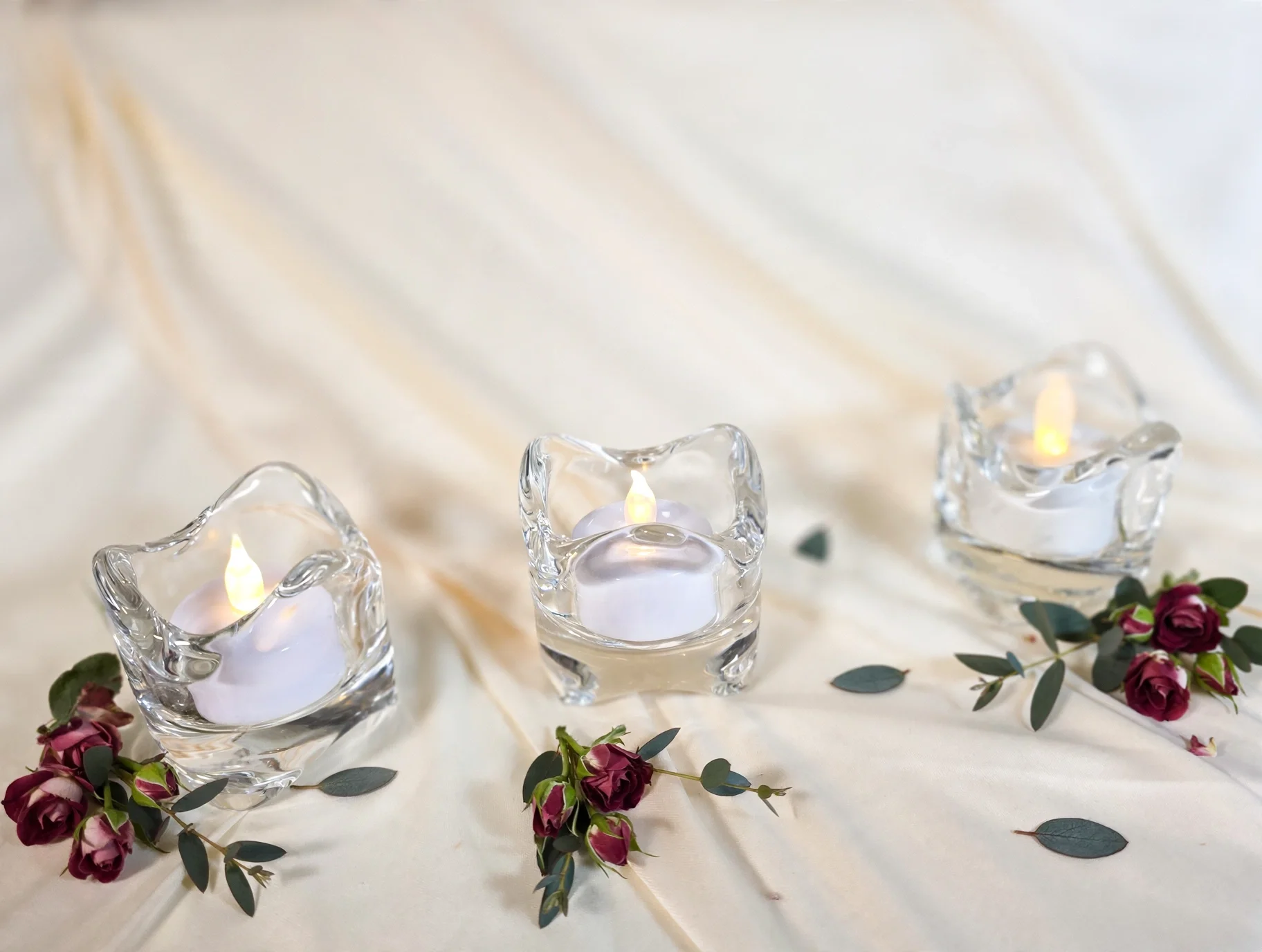 Three sculpted glass candle holders with glowing LED candles on cream satin fabric