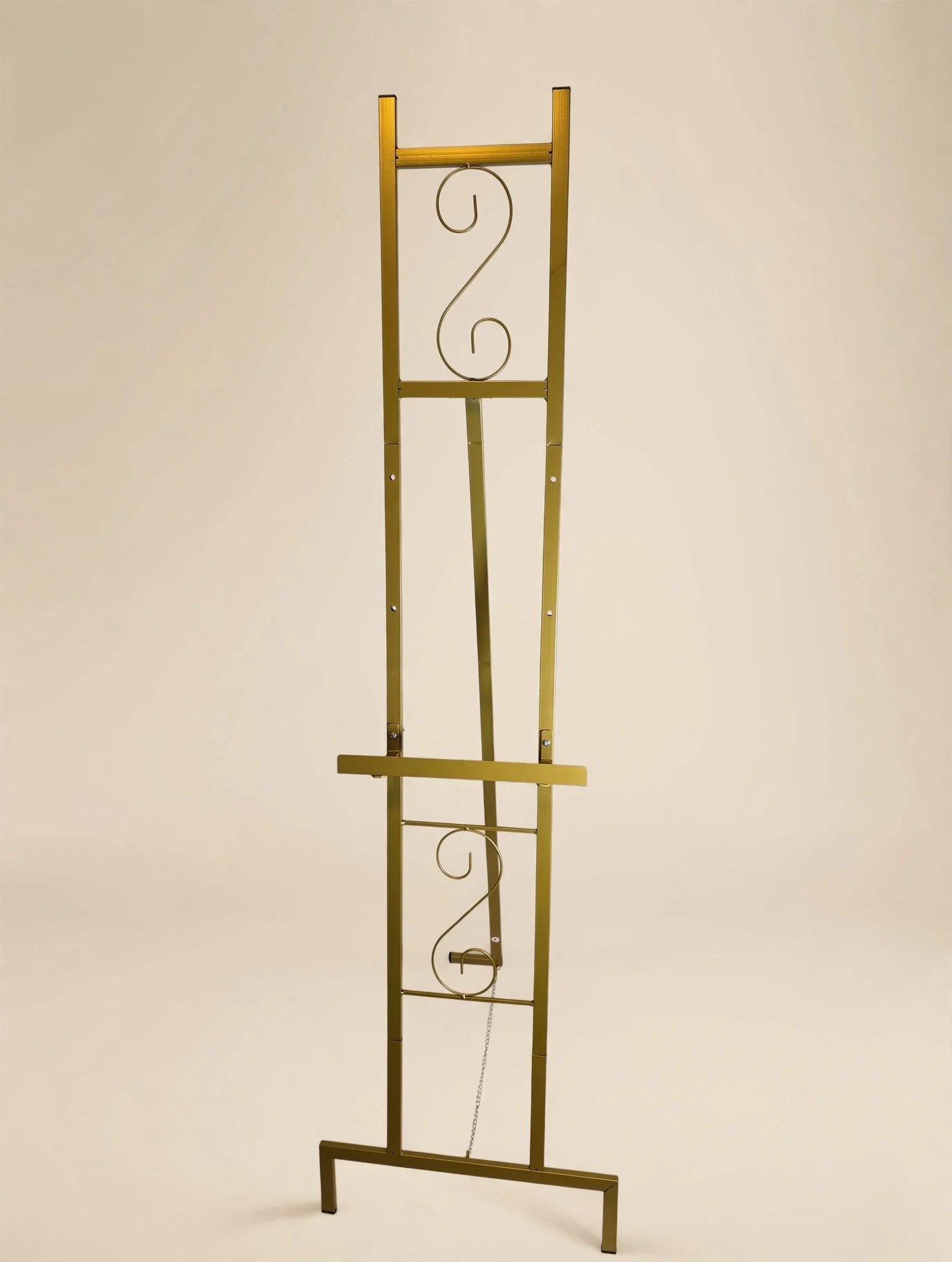 Tall gold metal easel with decorative scroll design and chain support against beige background