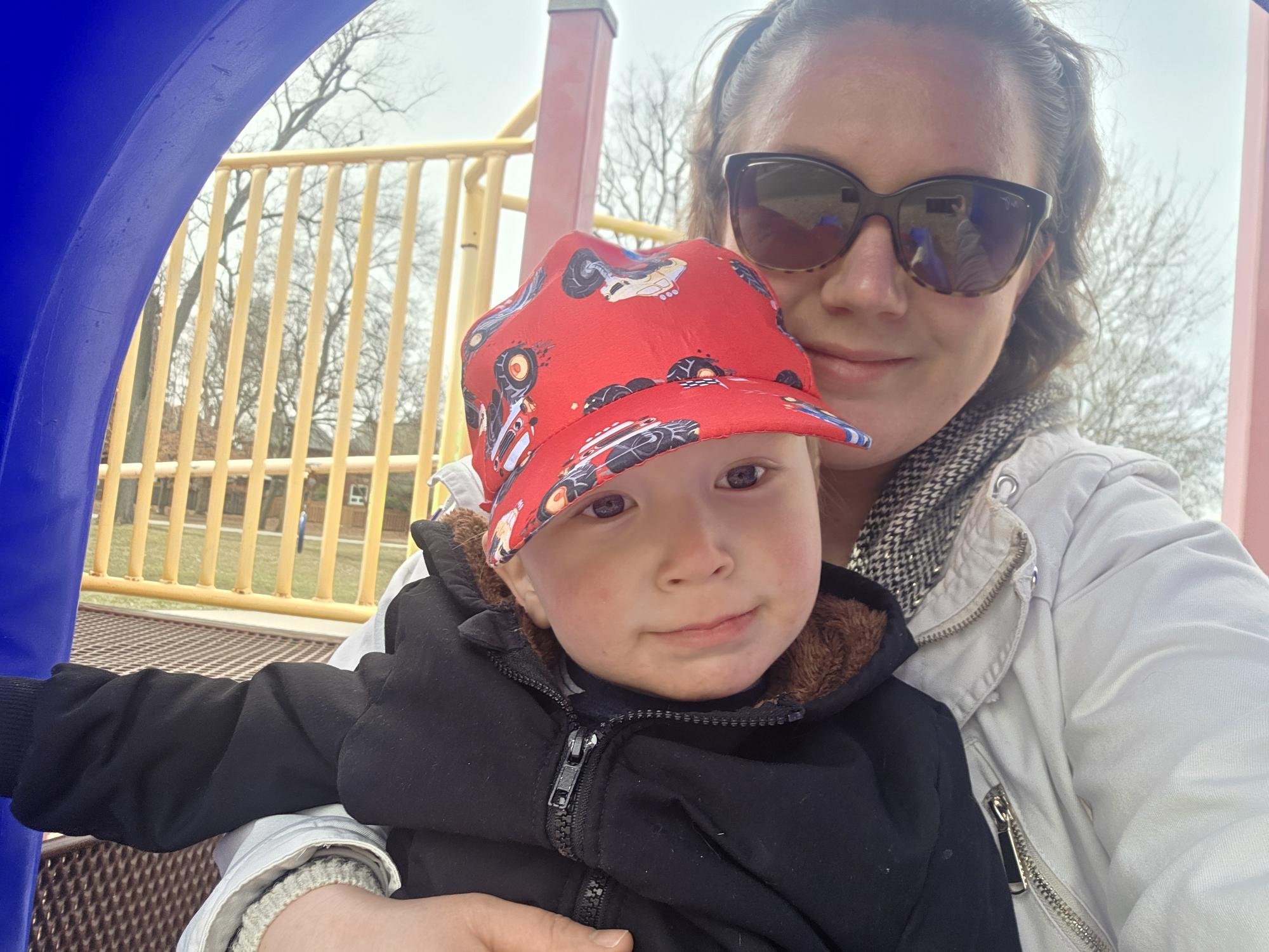 Adult woman with curly hair wearing sunglasses and a light-colored jacket, smiling while taking a selfie with toddler boy in a cap with cartoon vehicles at playground.