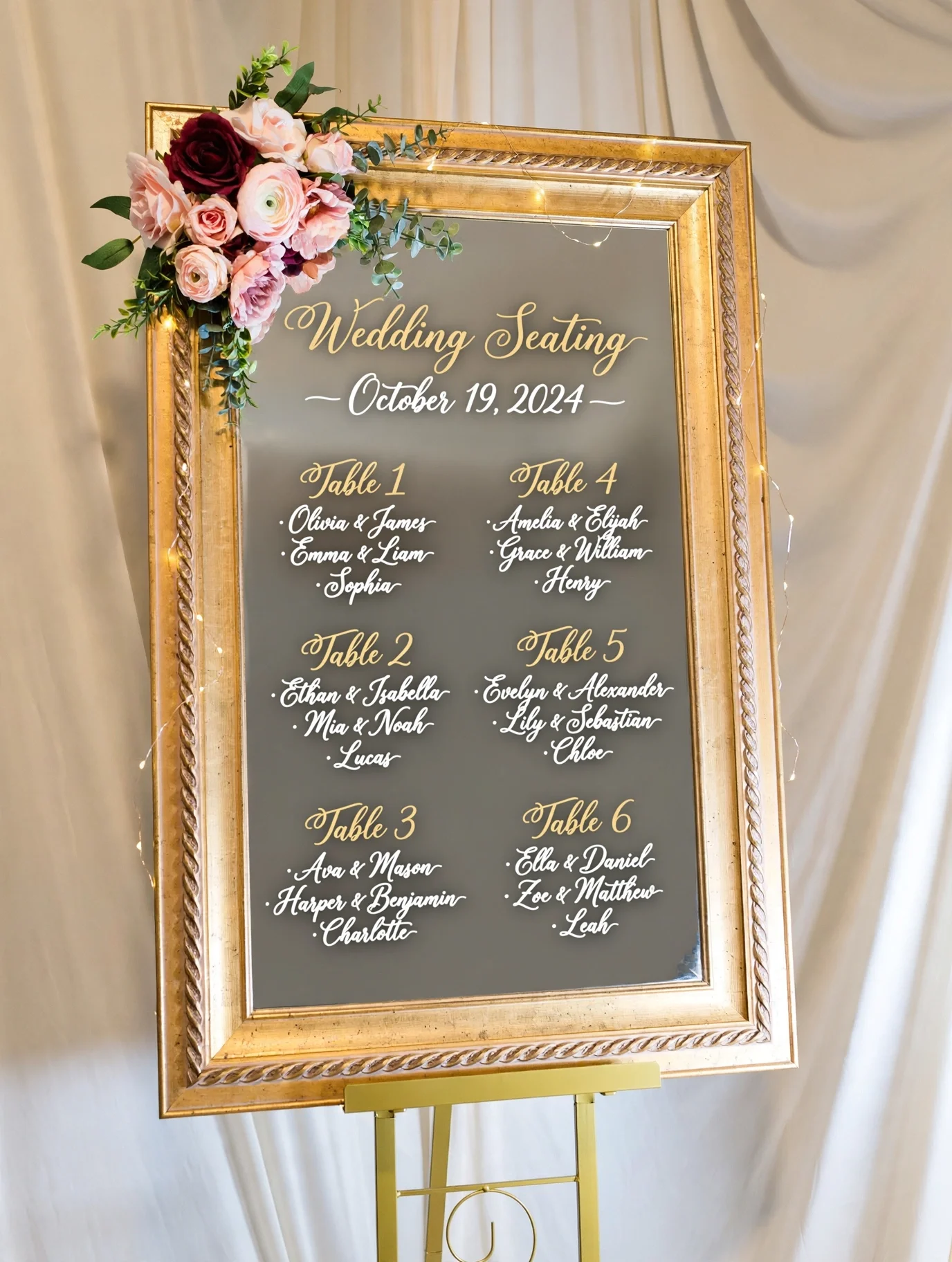 Gold framed mirror seating chart with floral accents displayed against soft draped fabric