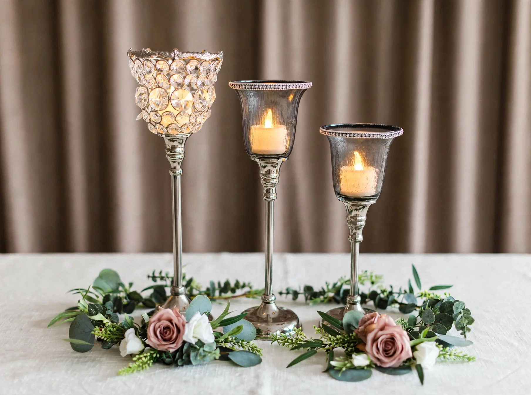 Three glass candle holders with glowing candles, roses, and greenery accents