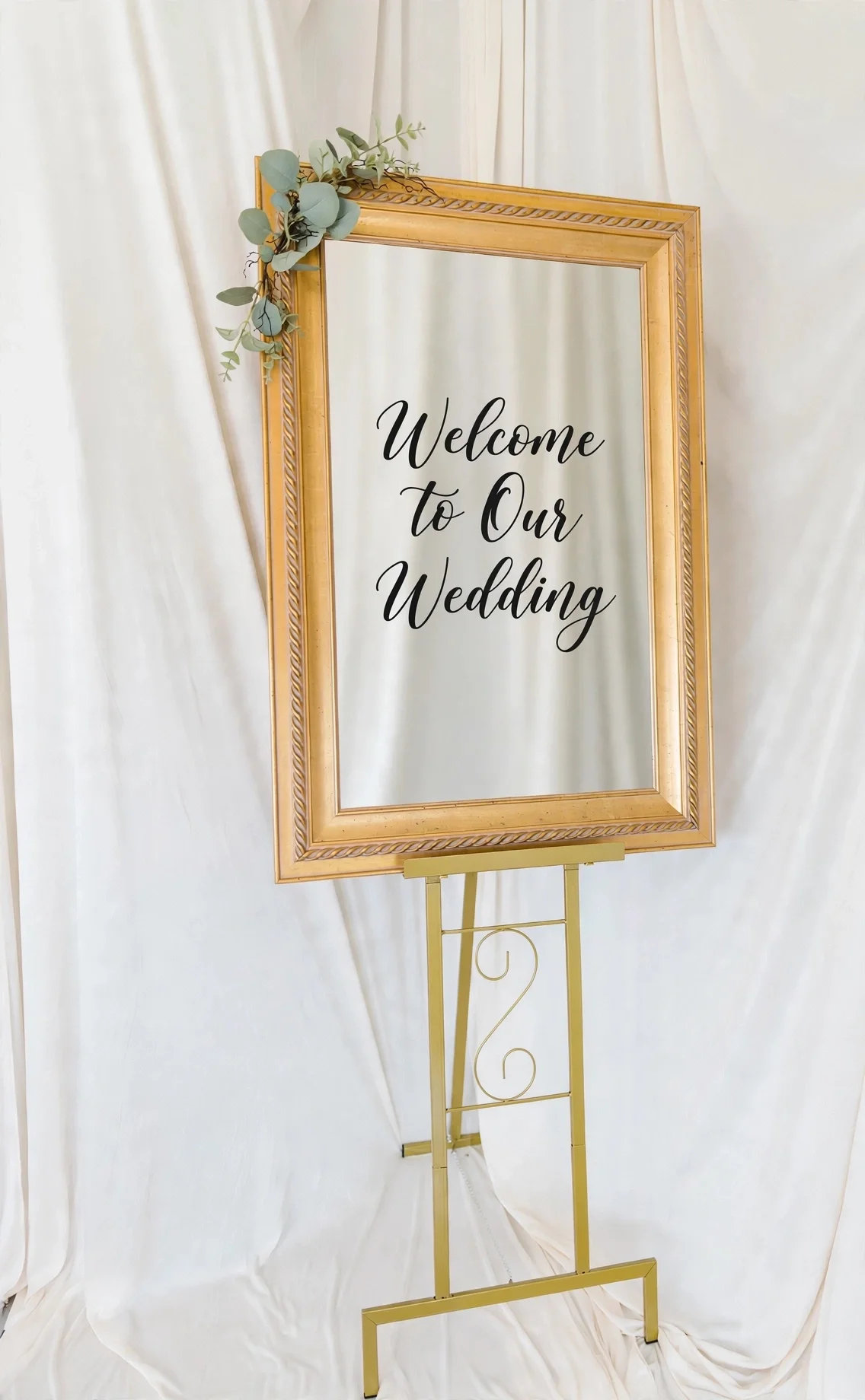 Gold framed welcome sign with eucalyptus greenery on a gold stand in front of white draping
