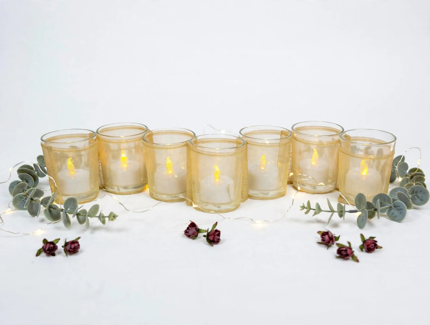 Seven glass candle holders wrapped in gold mesh with glowing LED candles on white fabric