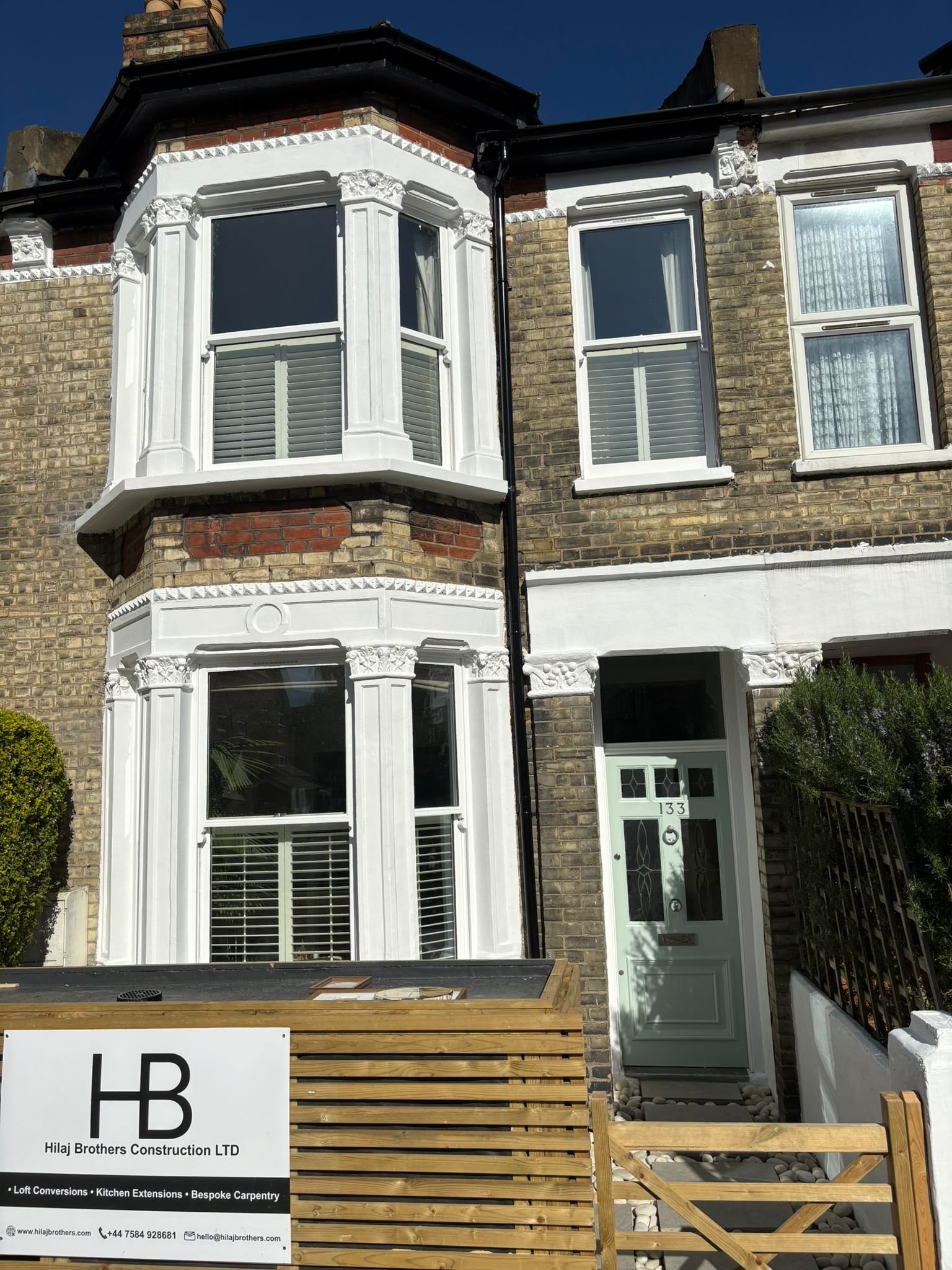 A multi-story brick house with white decorative trim around the windows and door, and a black gutter system. There is a sign in front indicating construction work by Hilaj Brothers Construction LTD.