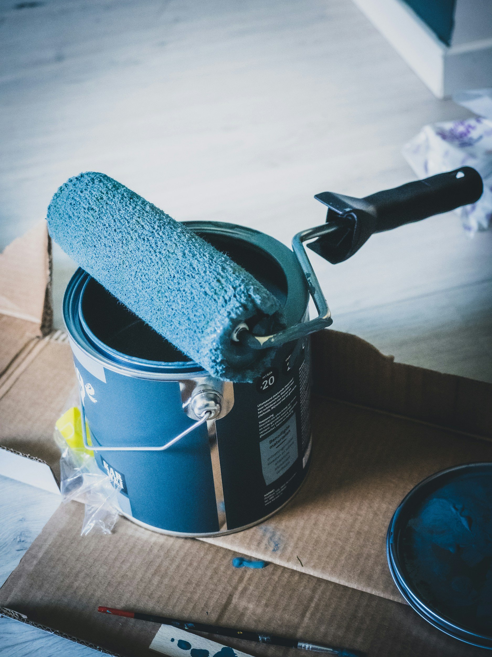 The image shows a paint roller with blue paint resting on the edge of an open can of blue paint. The can is placed on a box, and there are some plastic bags and a paintbrush nearby.