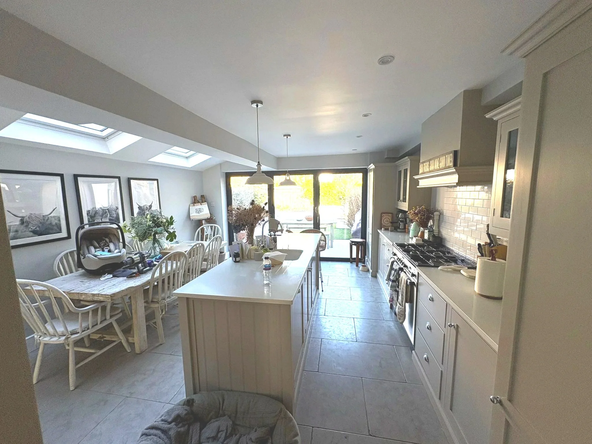 A bright kitchen and dining area with white cabinets, a large island, and a dining table with chairs. Skylights and large glass doors lead outside, and the room is decorated with framed artwork and plants.