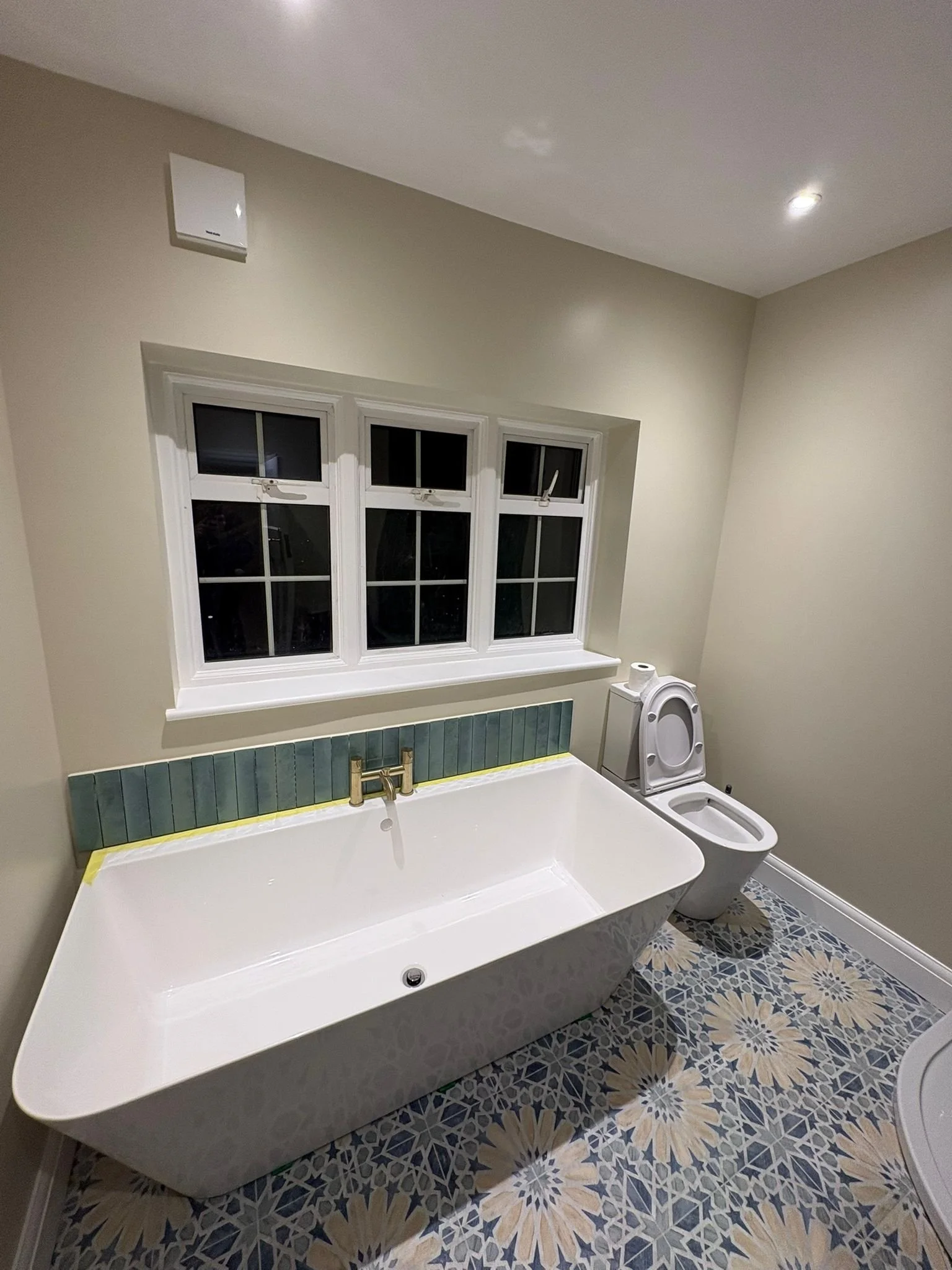 Bathroom with a white bathtub, a window above the tub, a toilet on the right, and patterned floor tiles.