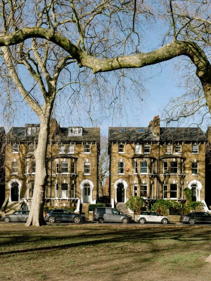 Three-story brick residential buildings with bay windows and steps leading to front doors, parked cars, a leafless tree in the foreground, and clear blue sky.